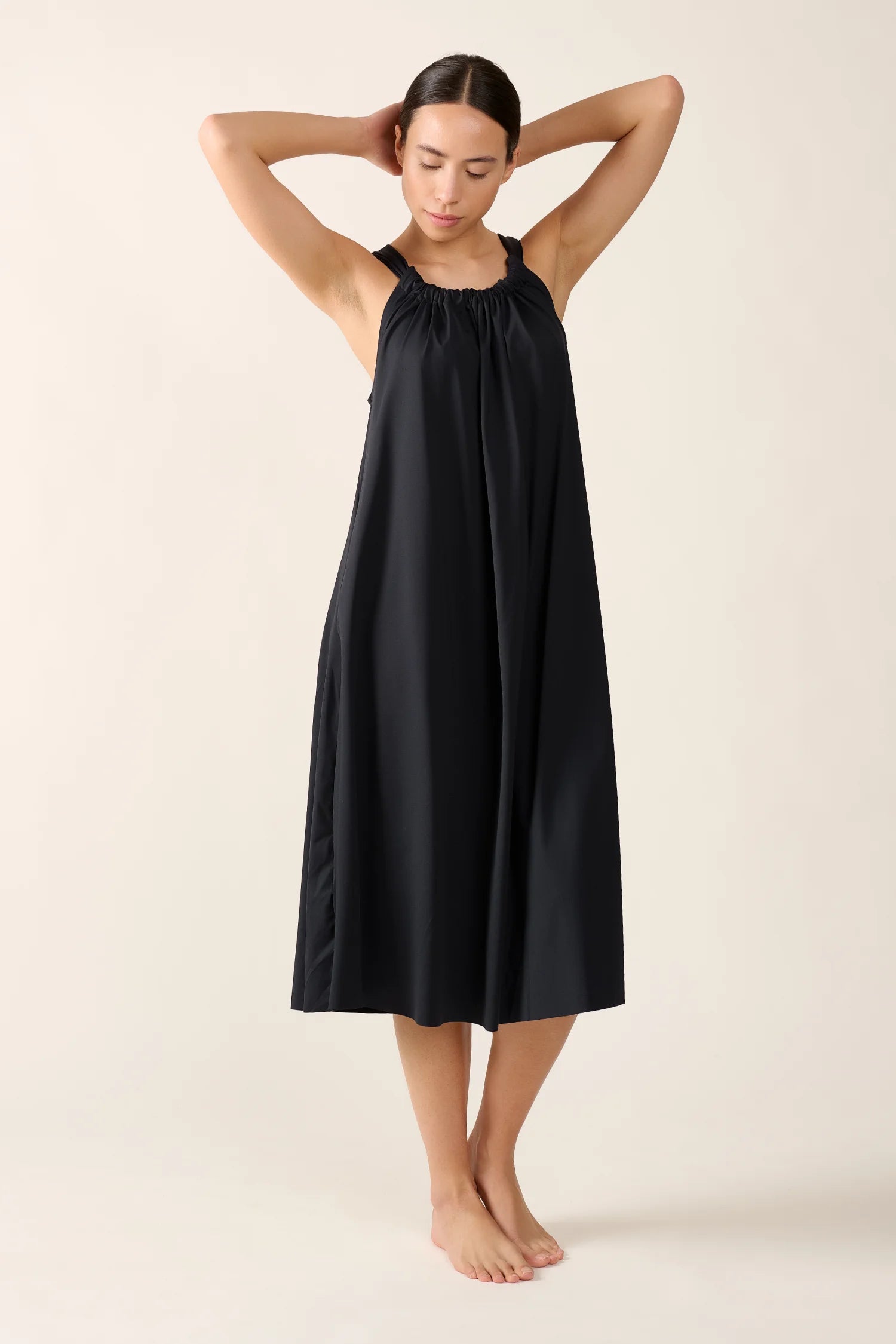 Frau in schwarzem MYMARINI-Kleid mit breiten Trägern, stehend, Arme hinter dem Kopf verschränkt.#color_black