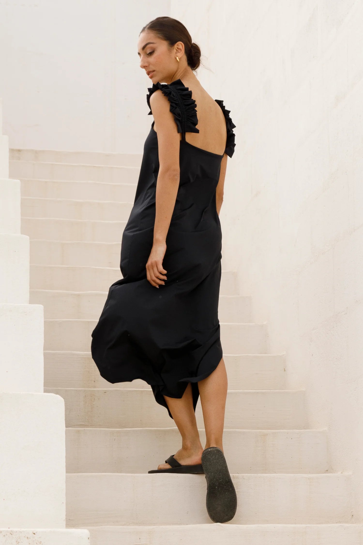 Schwarzes Kleid von MYMARINI mit schmalen Trägern und tiefem Rückenausschnitt, getragen von einer Frau auf einer Treppe.#color_black