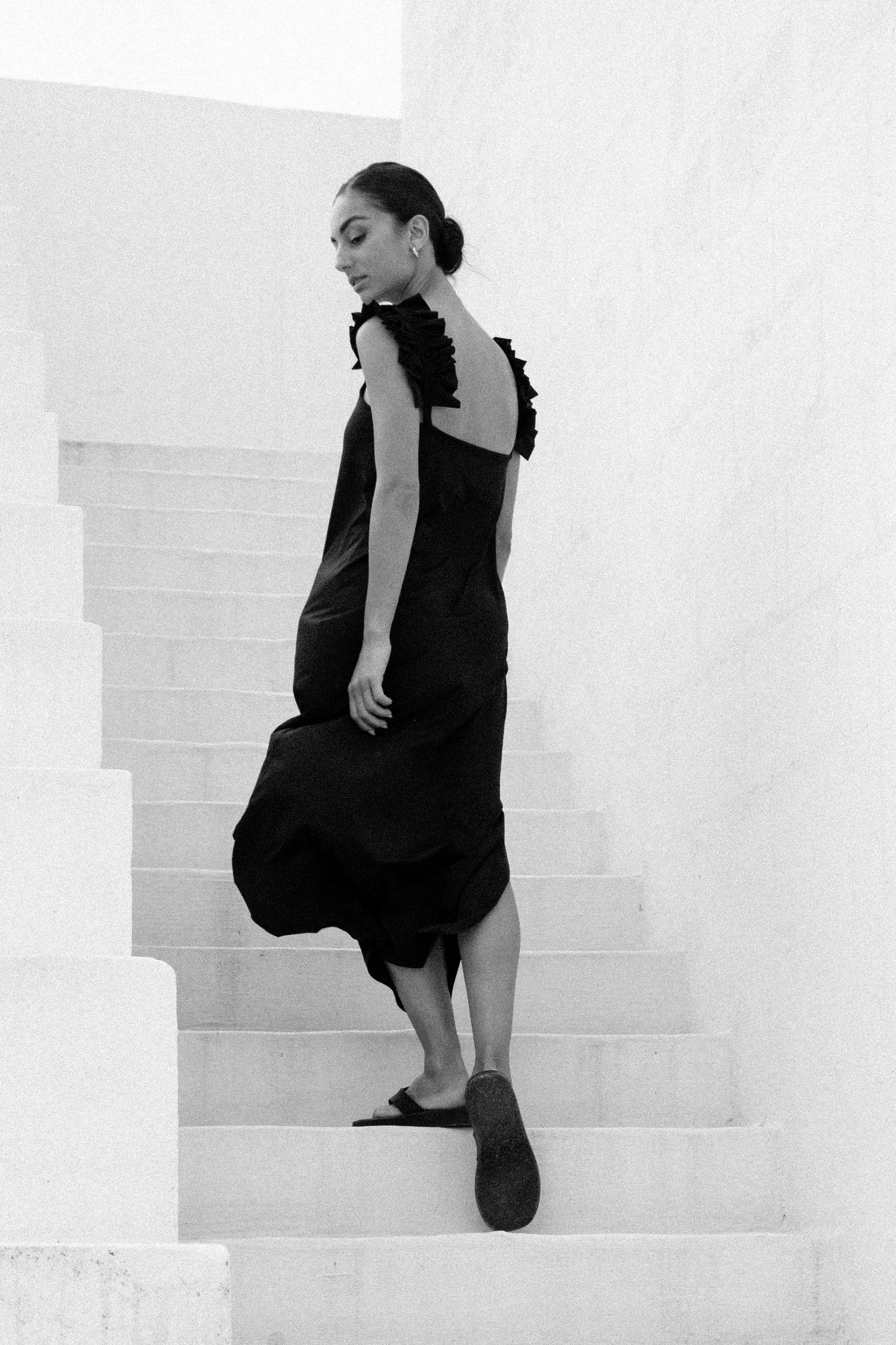 Schwarzes Kleid von MYMARINI mit schmalen Trägern und tiefem Rückenausschnitt, getragen von einer Frau auf einer Treppe.#color_black