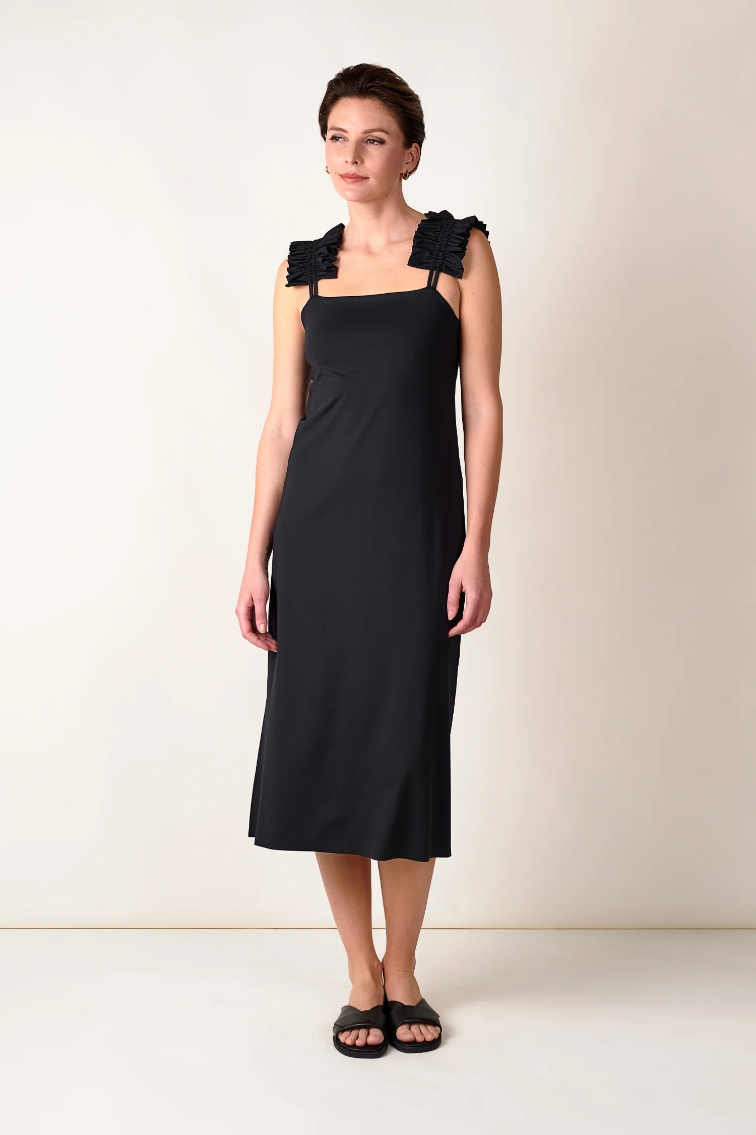 Frau trägt schwarzes MYMARINI-Kleid mit breiten Trägern, knielang, vor neutralem Hintergrund.#color_black