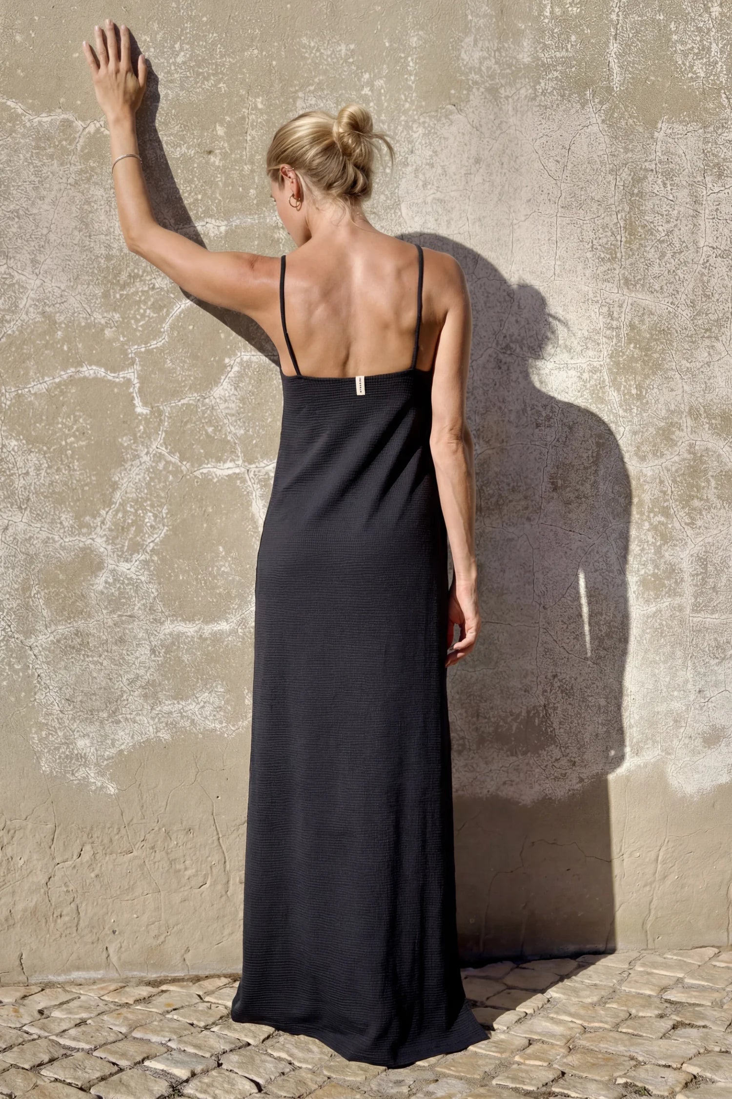 MYMARINI One-Piece Badeanzug mit Spaghettiträgern und offenem Rücken; Frau steht seitlich an einer Wand, Arm ausgestreckt.