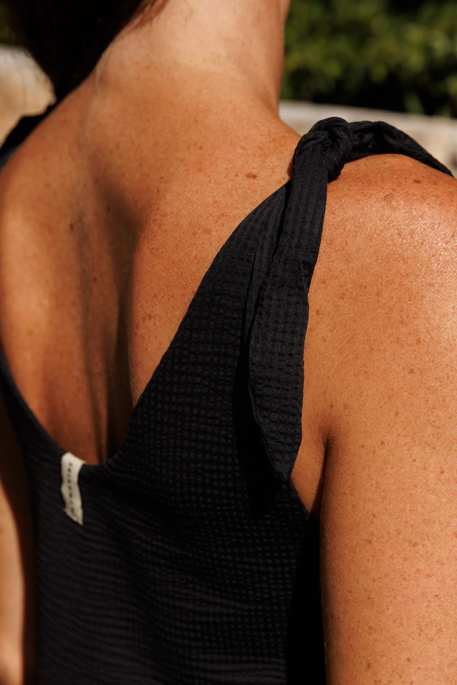 MYMARINI One-Piece mit gekreuzten Trägern im Rücken, schwarzer Bademodestoff in Nahaufnahme von Schulter und Oberarm.