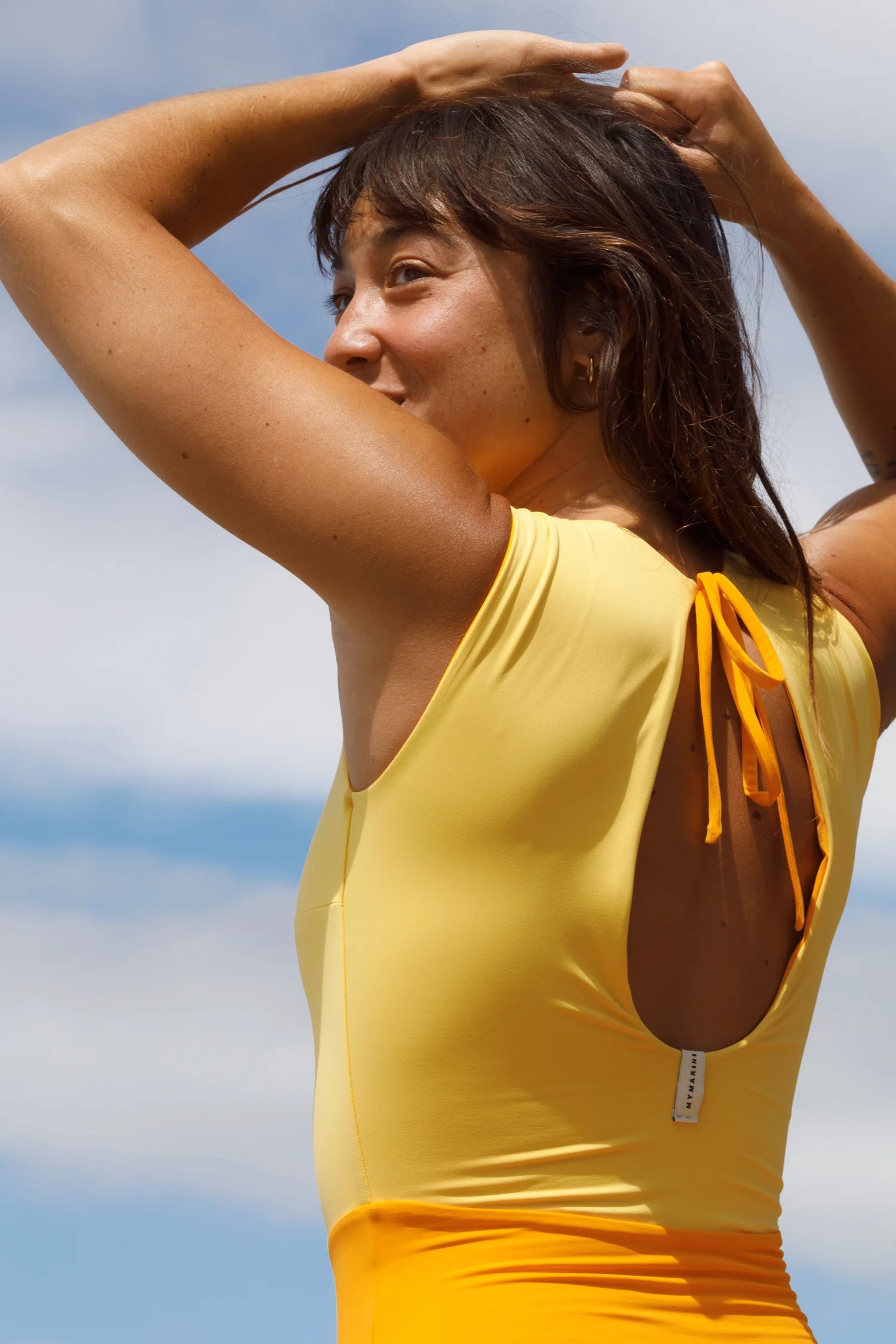 Frau in gelbem MYMARINI-Badeanzug mit tiefem Rückenausschnitt vor blauem Himmel.#color_buttermilk-soleil