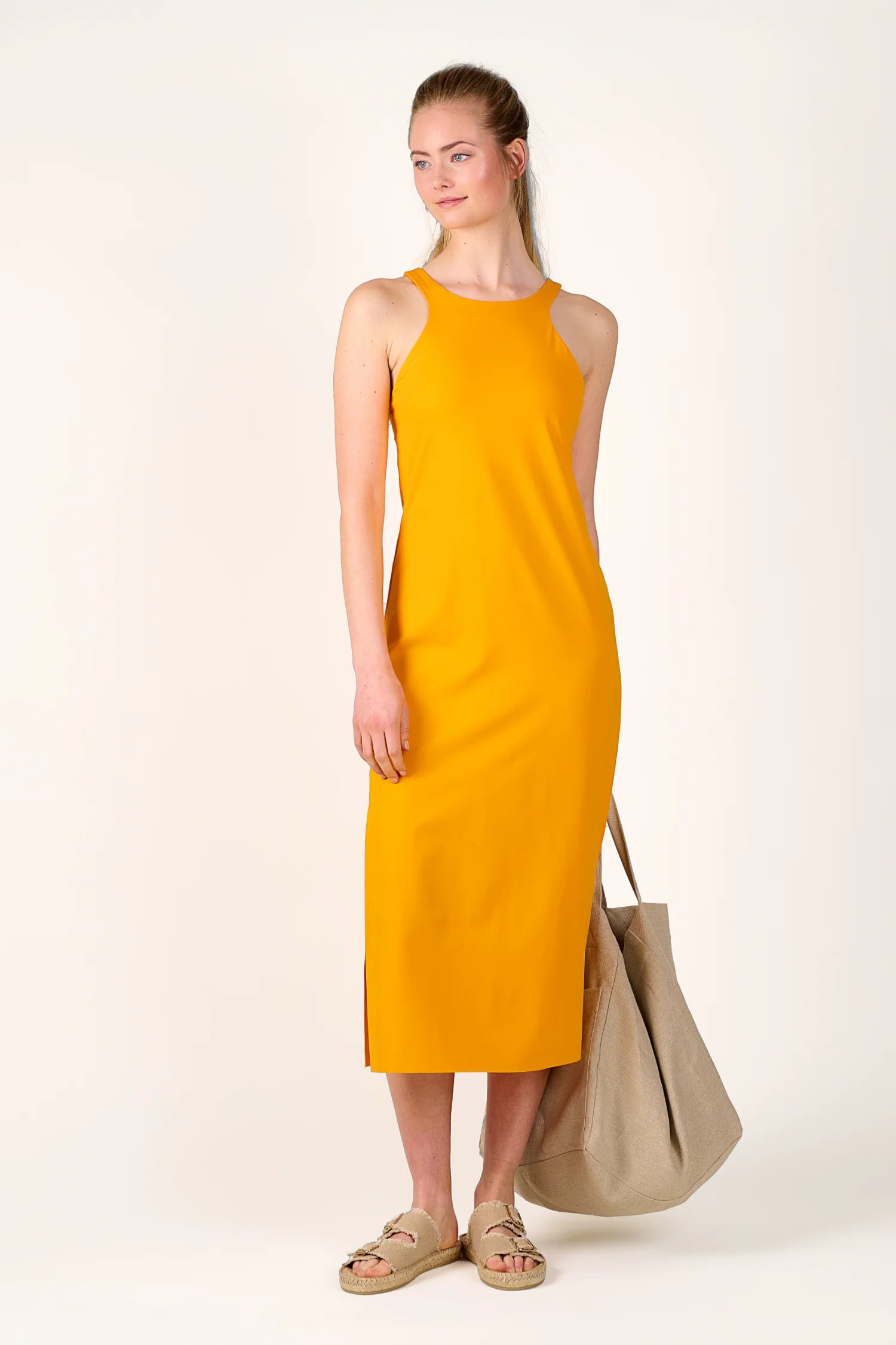 Frau in gelbem MYMARINI-Kleid mit Rundhalsausschnitt, hält eine große beigefarbene Tasche, trägt Sandalen.#color_buttermilk-soleil