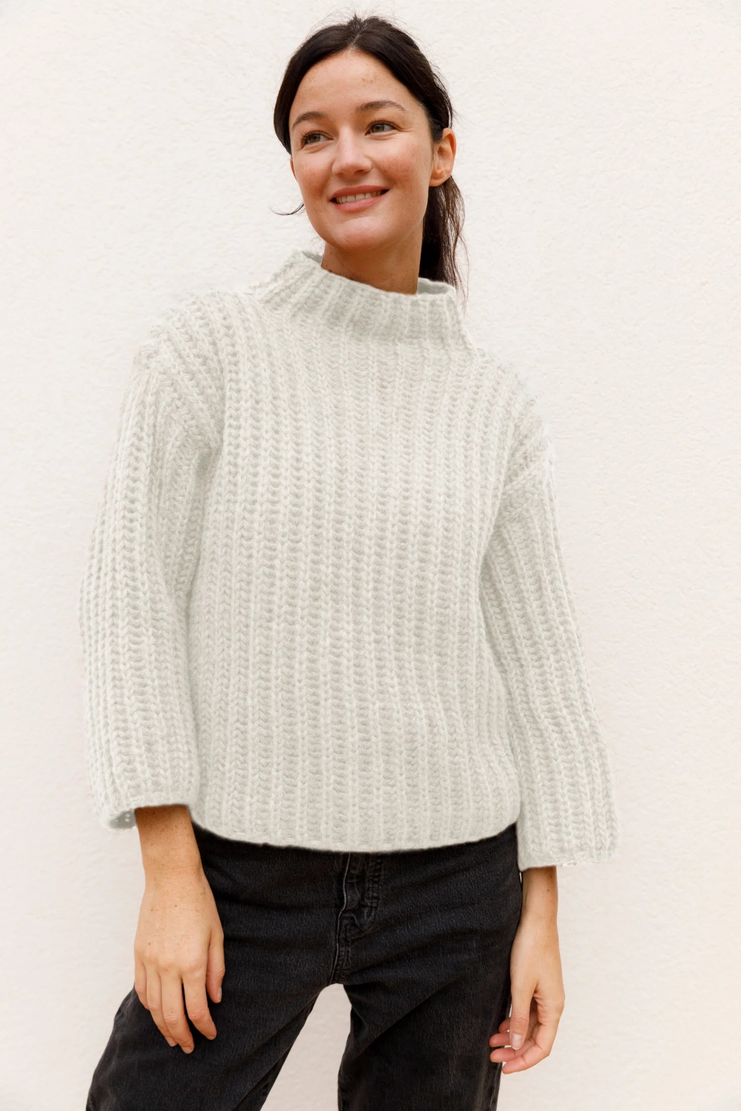 Frau in cremefarbenem Strickpullover mit Stehkragen und schwarzen Jeans vor hellem Hintergrund.#color_white-stone