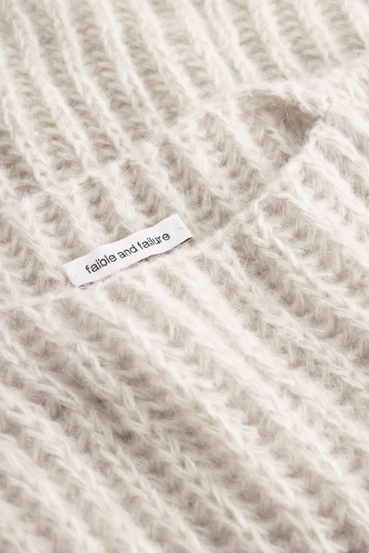 Nahaufnahme eines cremefarbenen Strickpullovers mit sichtbarem Label "fable and failure".#color_white-stone