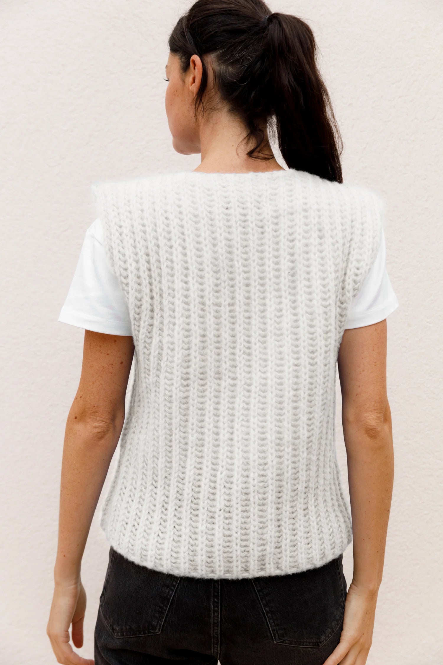 Frau in weißem T-Shirt und gestrickter Weste von MYMARINI, von hinten, vor hellem Hintergrund.#color_white-stone