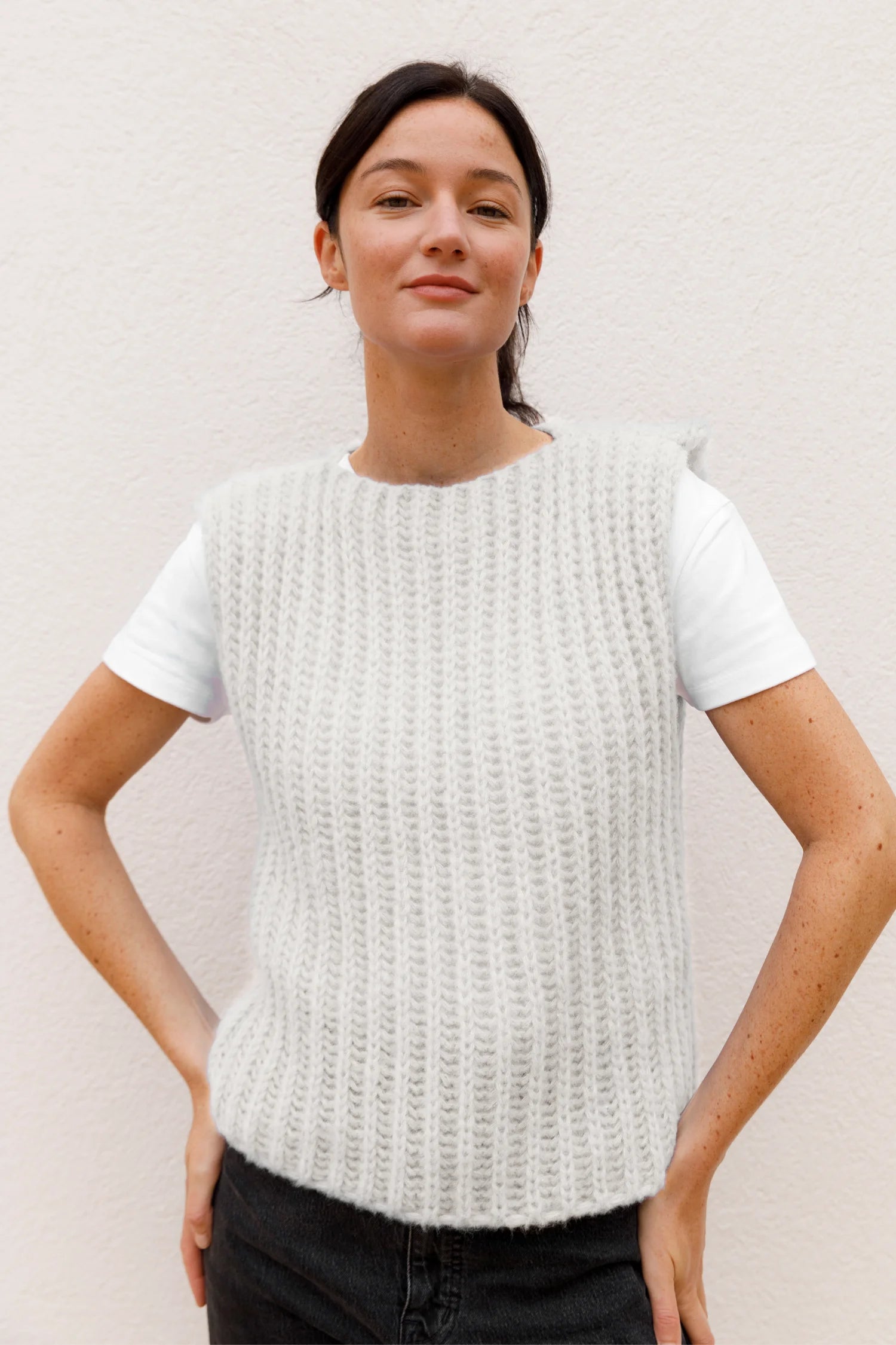 Frau trägt weißen Strick-Top über weißem T-Shirt, lächelt leicht, vor hellem Hintergrund.#color_white-stone