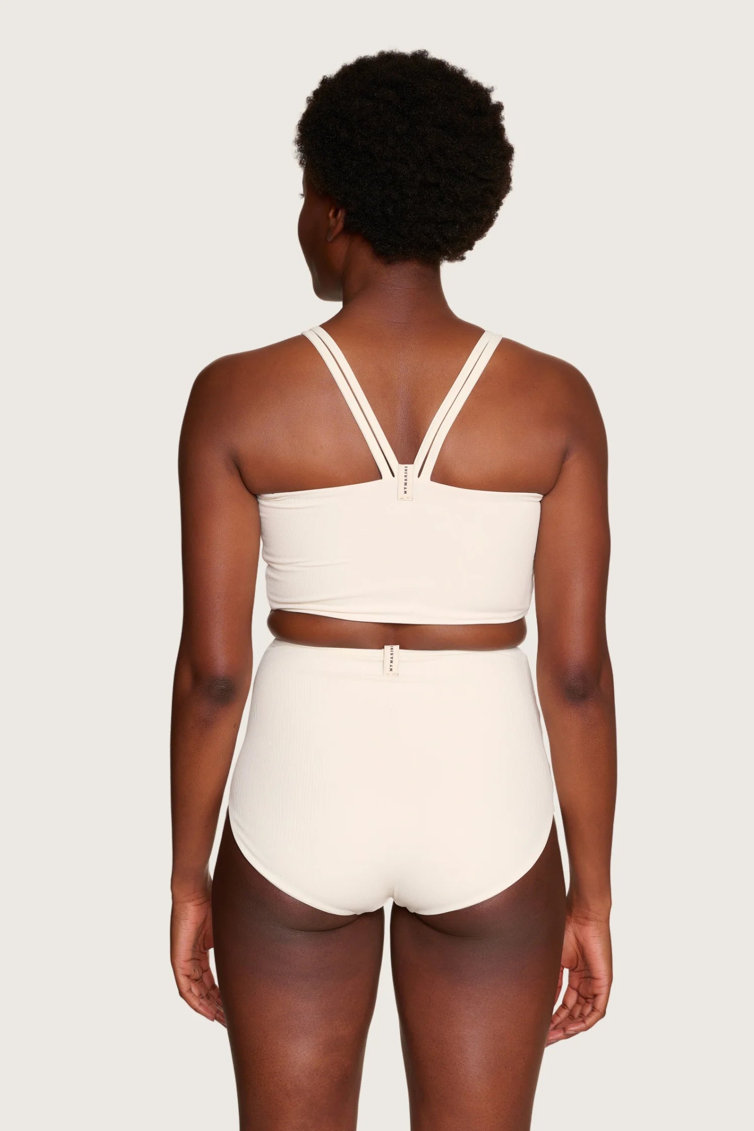 Rückansicht einer Person in cremefarbenem MYMARINI-Bikinitop mit breiten Trägern und High-Waist-Bikinihose.#color_off-white