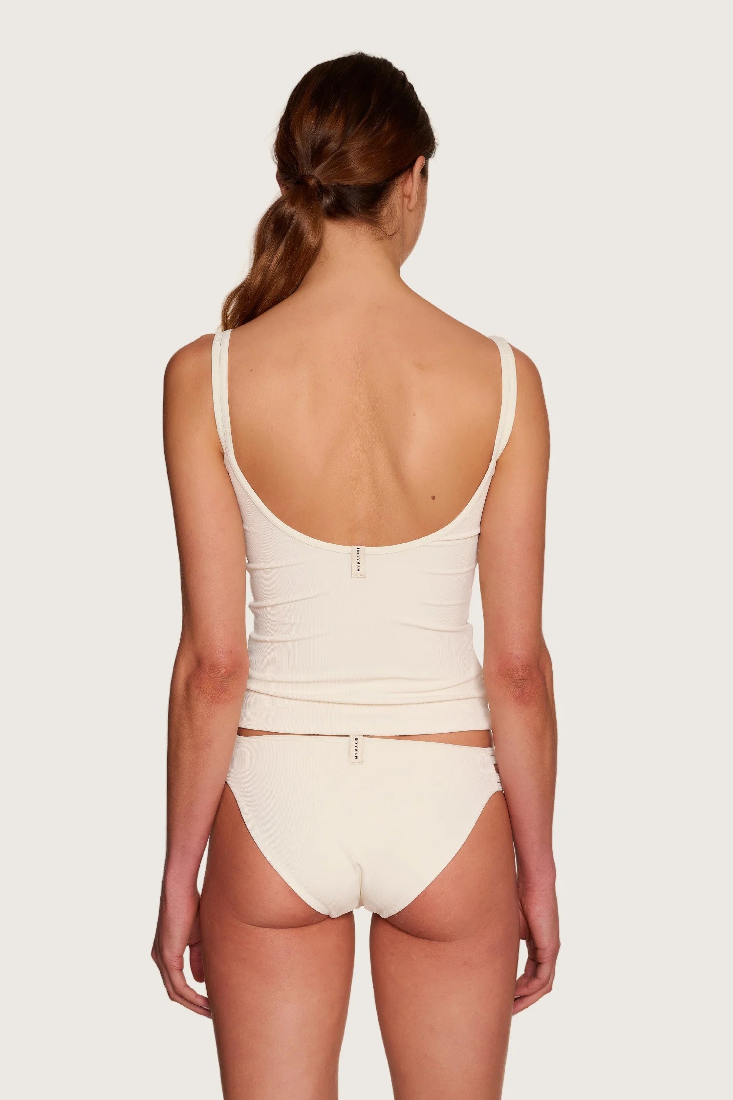 Rückansicht einer Person in cremefarbenem MYMARINI-Top mit tiefem Rückenausschnitt und passender Bikinihose, hoher Beinausschnitt.#color_off-white