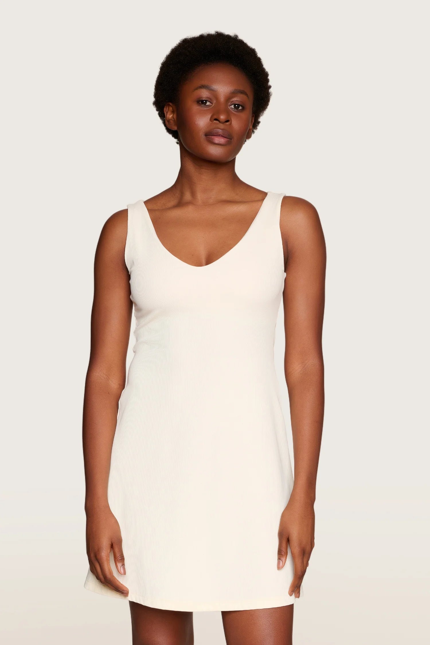 Person trägt ein ärmelloses, cremefarbenes MYMARINI-Kleid mit V-Ausschnitt vor neutralem Hintergrund.#color_off-white