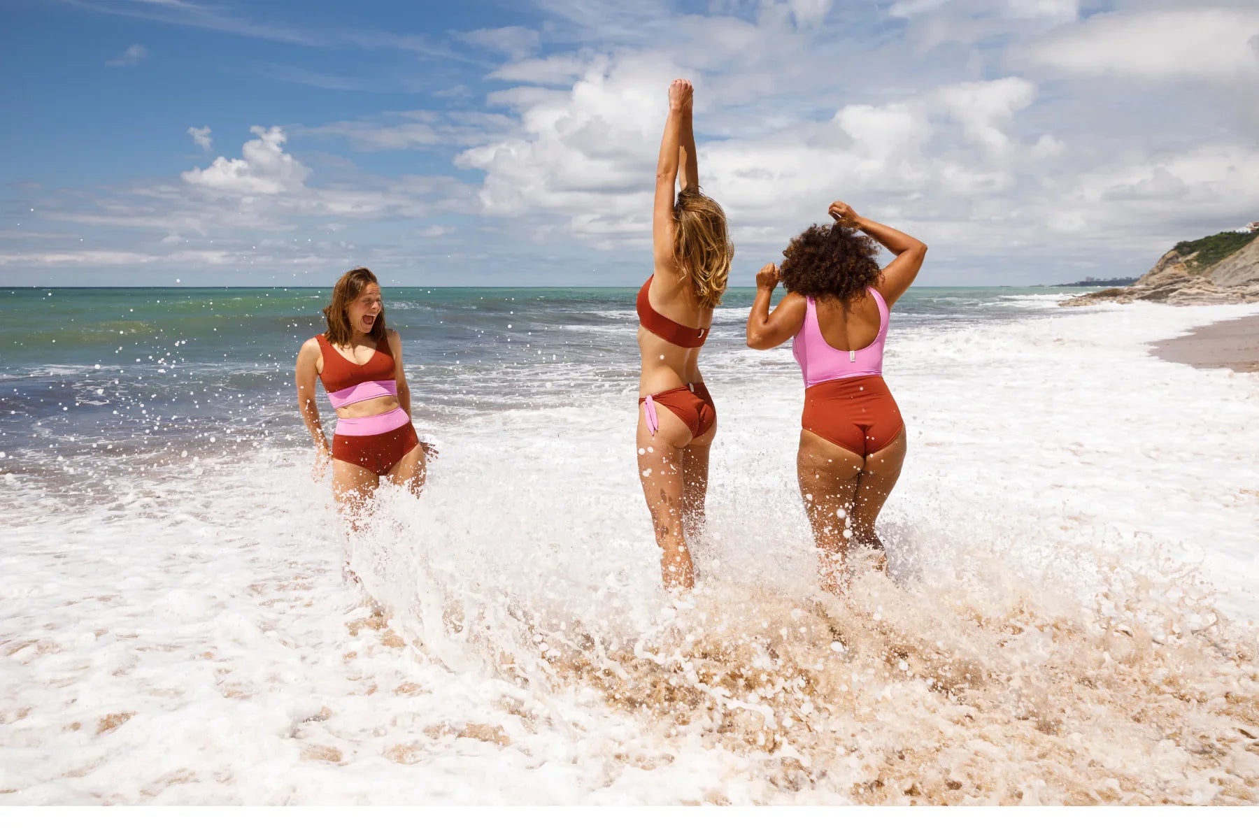 Drei Frauen in farbenfroher MYMARINI Swimwear spielen fröhlich in den Wellen am Strand; ihre Badeanzüge und Bikinis in Rot und Rosa strahlen Lebensfreude und Vielfalt aus.