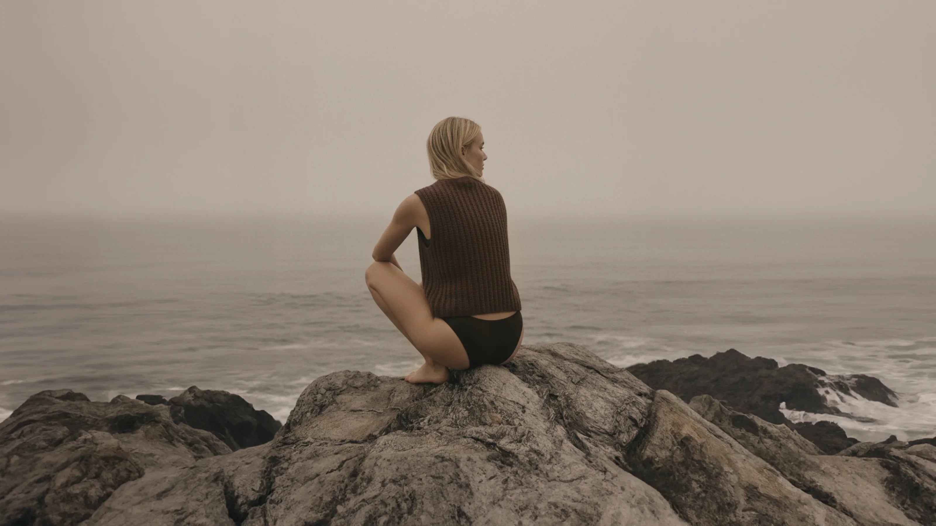 Frau hockt auf Felsen und schaut über das neblige Meer in der Ferne.