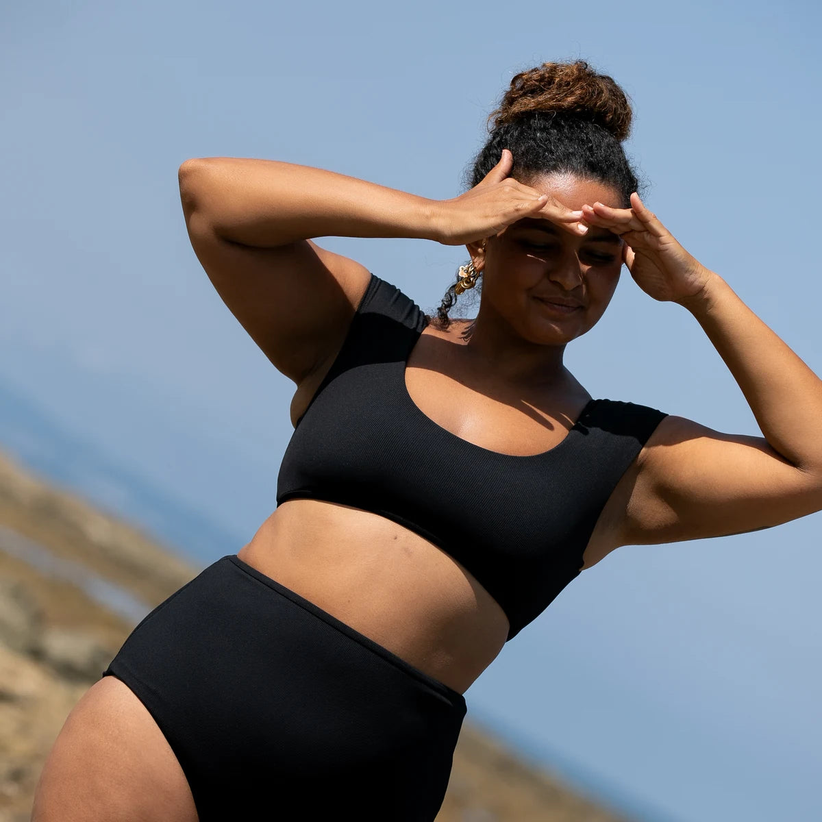 Woman shading her eyes with both hands in the black SWIM APPAREL bikini, blue sky behind her