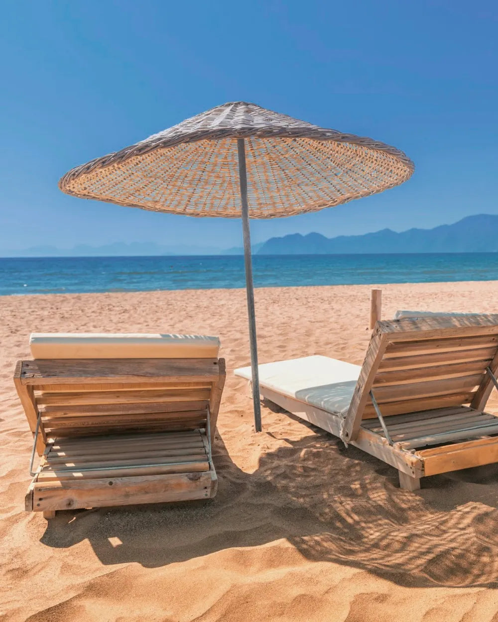 Sandstrand mit zwei Holz-Liegen, Sonnenschirm im Strohgeflecht und Blick auf Meer und ferne Berge.