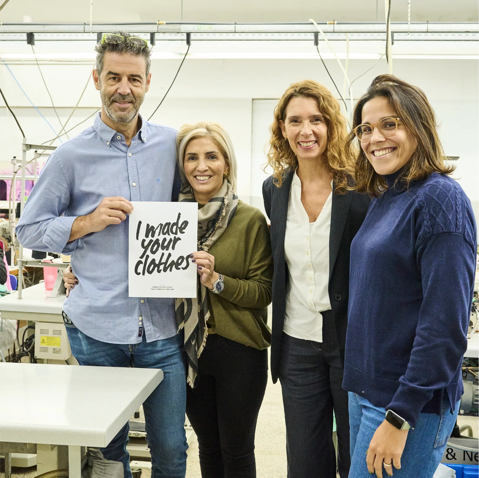 Vier Personen in einer Werkstatt, eine hält ein Schild mit der Aufschrift "I made your clothes".