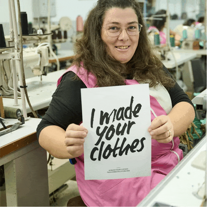 Frau in Näherei hält Schild mit der Aufschrift "I made your clothes", lächelnd in die Kamera blickend.