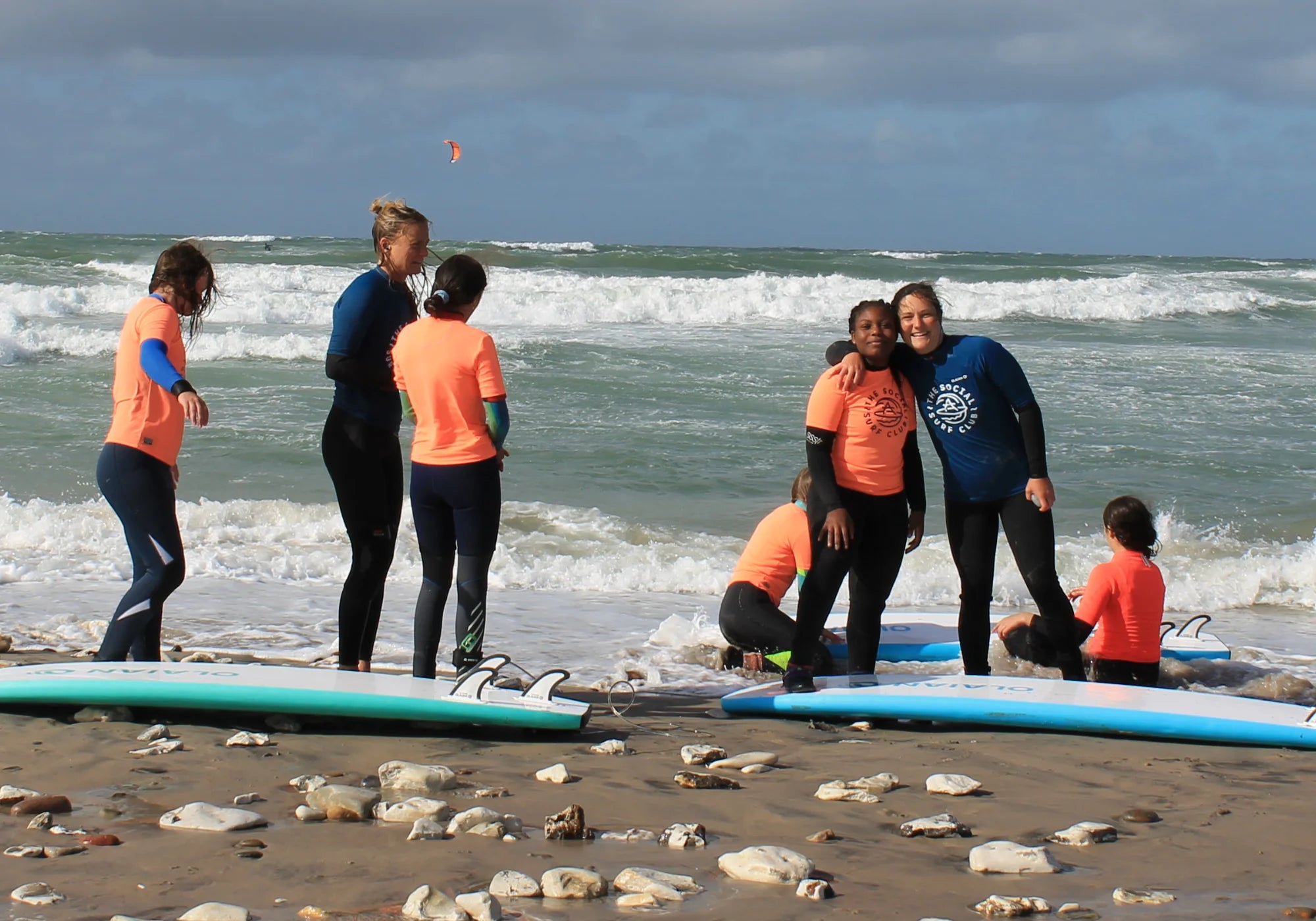 Menschen in Neoprenanzügen und MYMARINI-Shirts am Strand mit Surfbrettern, lächelnd und in Gruppen posierend.