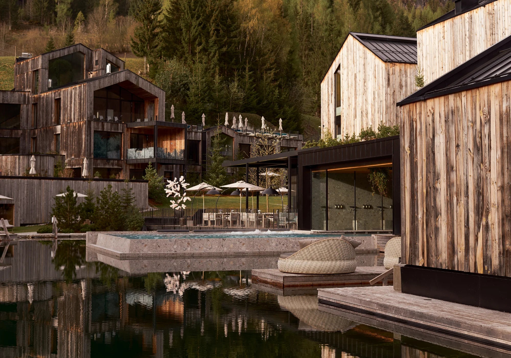 Moderne Holzhäuser und Pool in alpiner Landschaft, umgeben von Bäumen und Bergen.