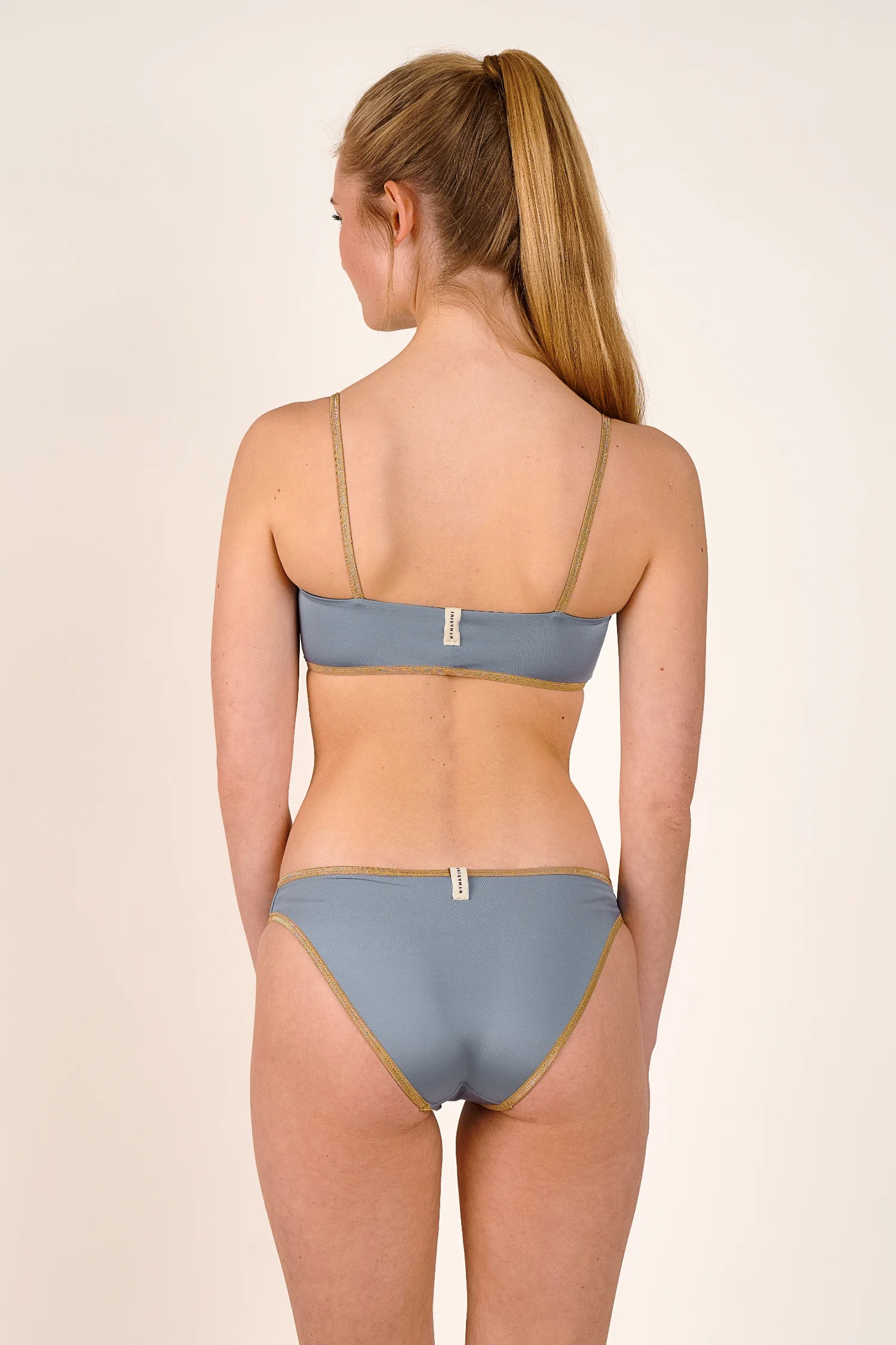 Rückansicht einer Frau in einem MYMARINI Bikini mit Spaghettiträgern und hohem Beinausschnitt in Blau.#color_crystal-grey