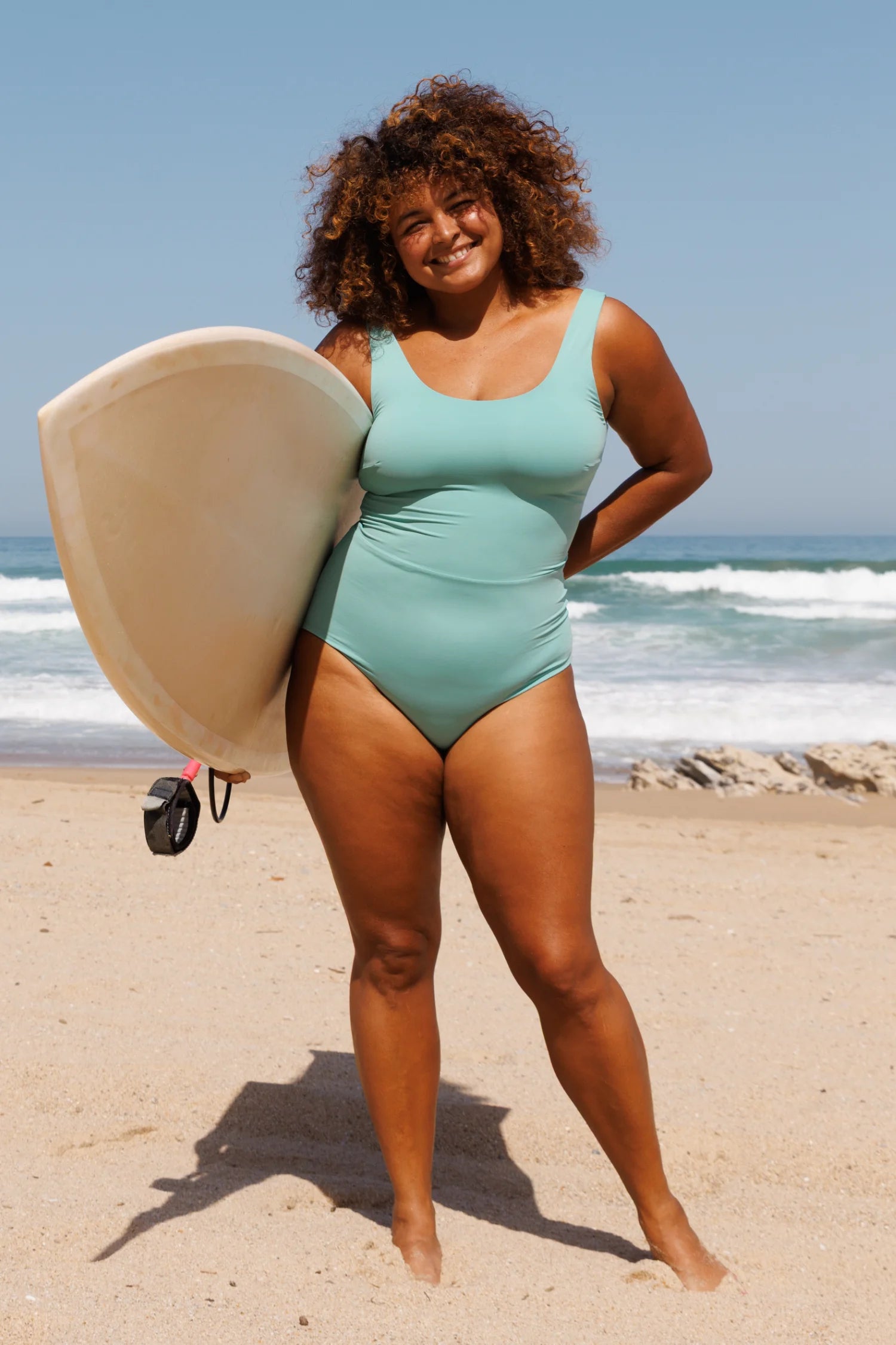 MYMARINI einteiliger Badeanzug mit breiten Trägern in Mint am Strand, mit Surfbrett, freundliche Pose.#color_oak-eucalyptus