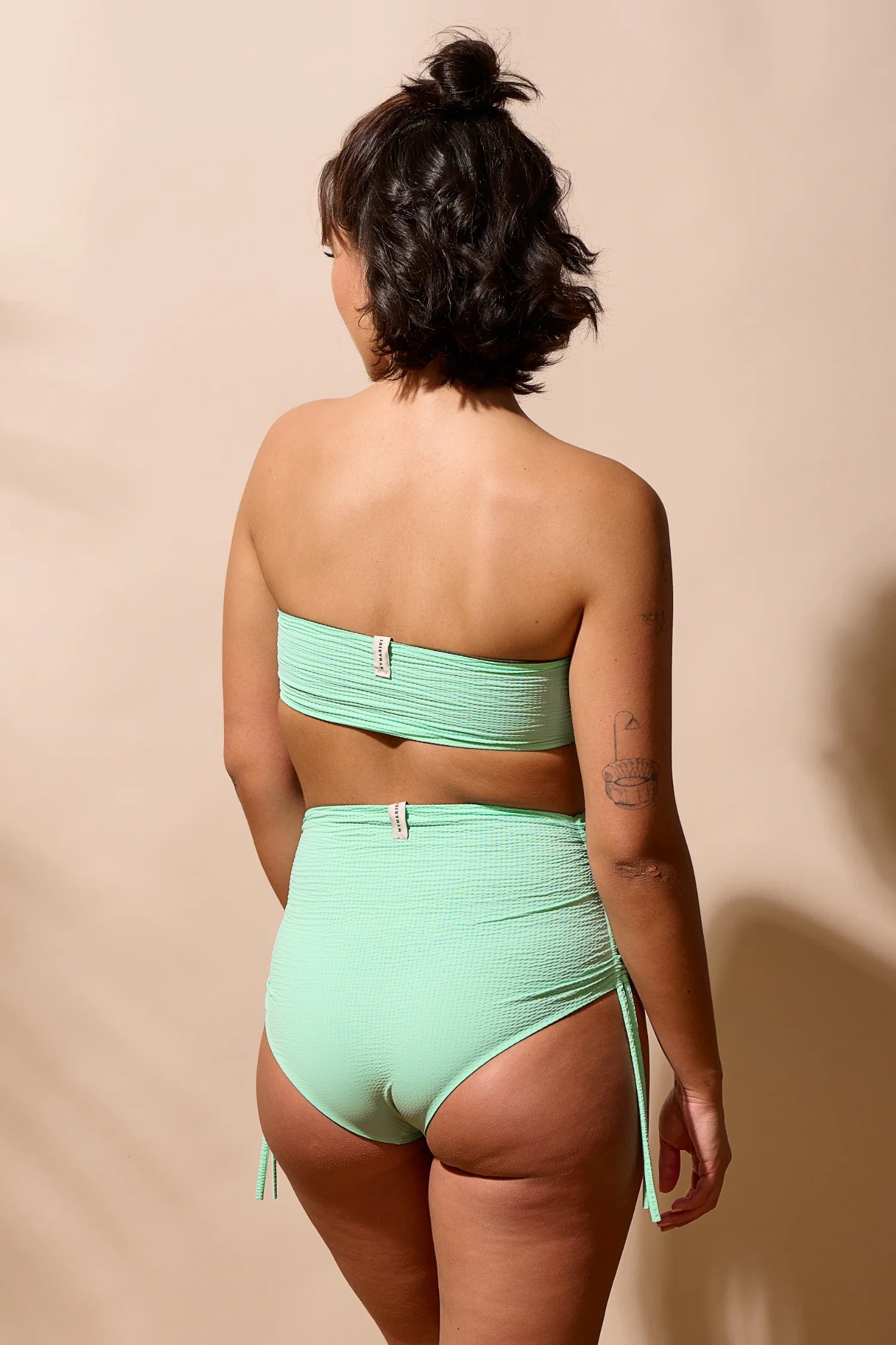 Frau trägt MYMARINI Bikini mit Bandeau-Top und High-Waist-Hose in Mintgrün, Rückansicht, vor beigem Hintergrund.#color_lagoon-seaweed