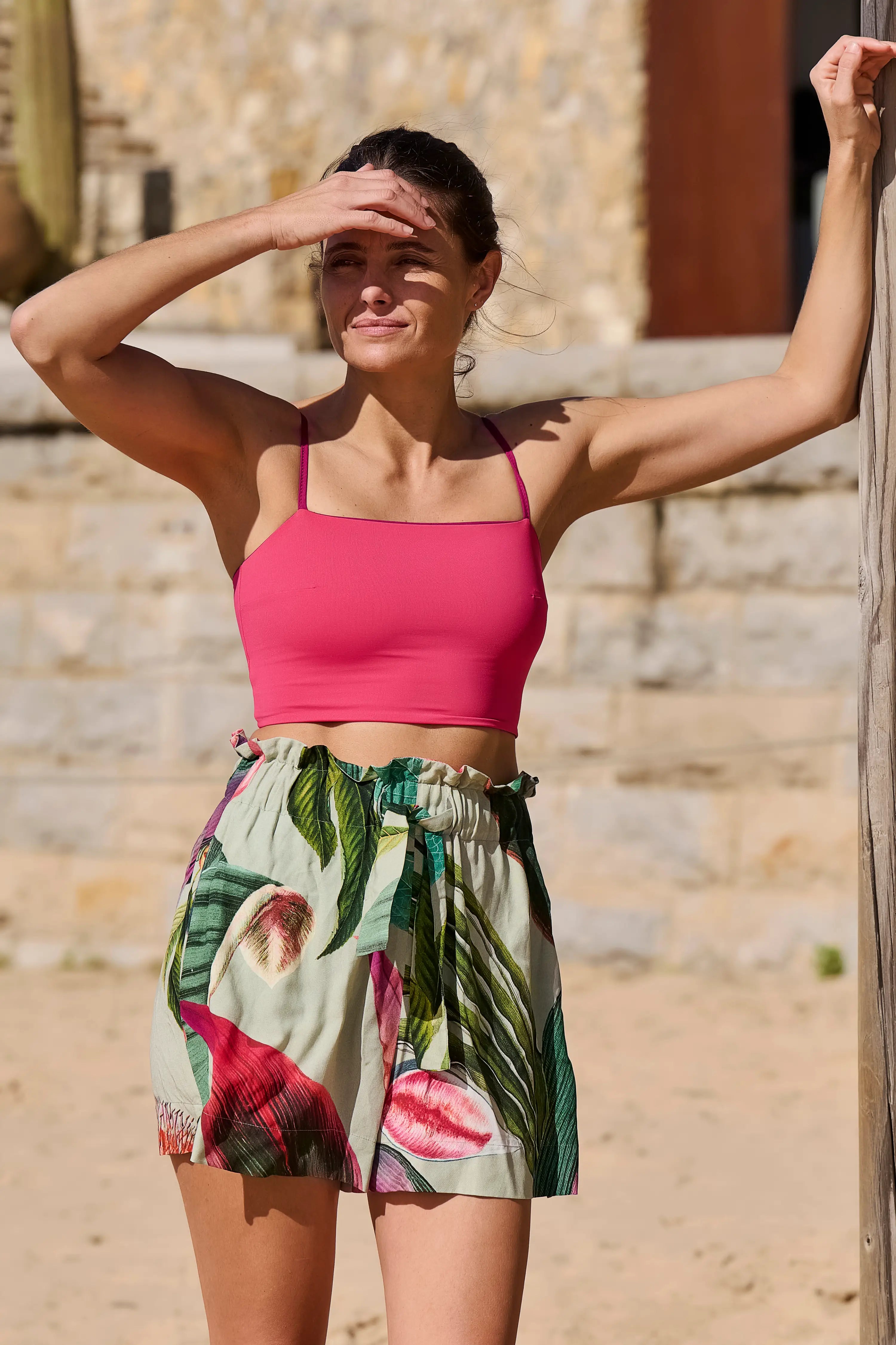 Frau am Strand in MYMARINI Spaghettiträger-Bikinitop und Shorts mit tropischem Muster, Hand über den Augen gegen die Sonne.#color_tropical-print