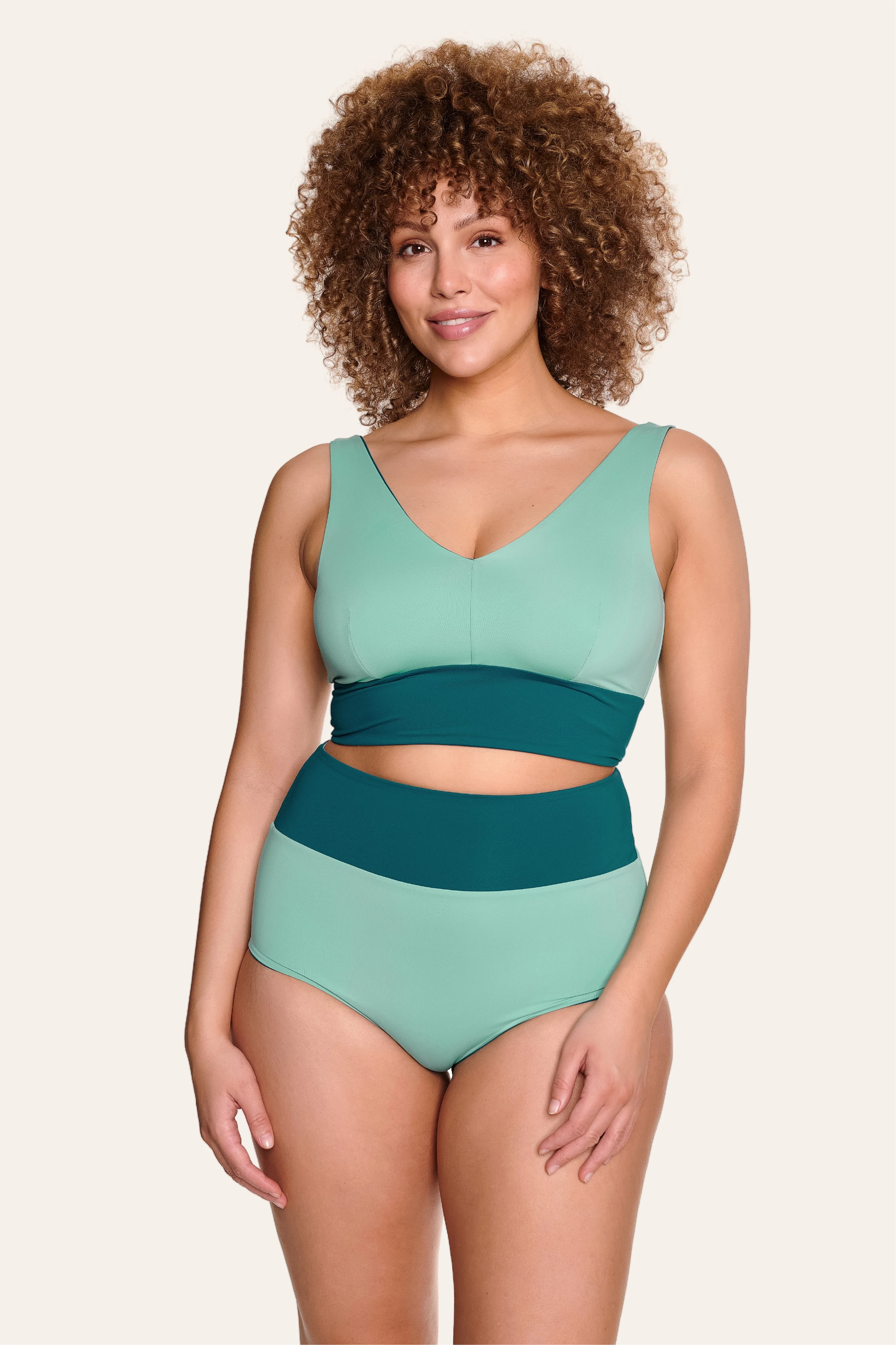 Frau trägt MYMARINI Bikini mit V-Ausschnitt und High Waist in Grün- und Blautönen, lächelnd vor hellem Hintergrund.#color_eucalyptus-petrol