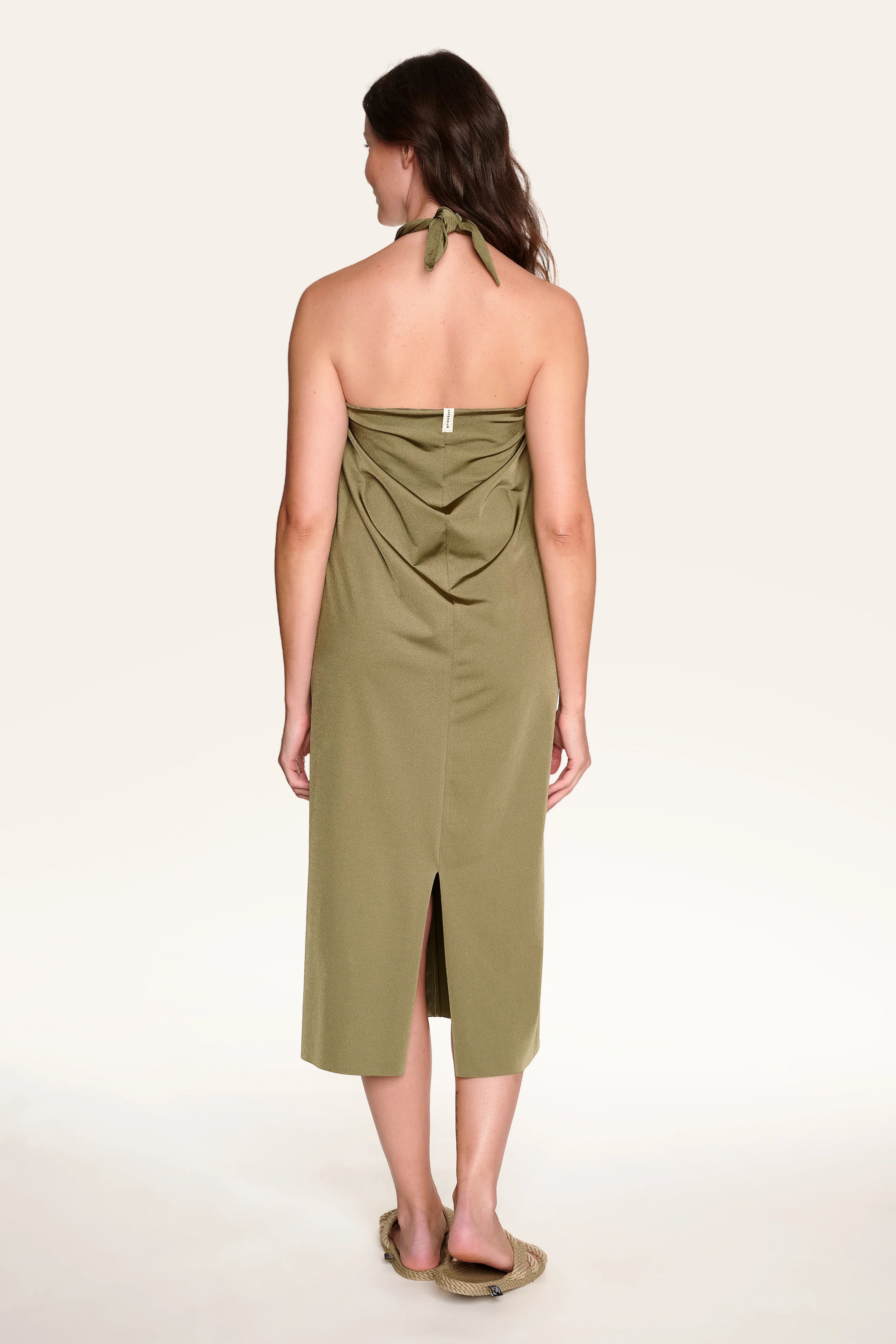 Rückansicht einer Frau in einem olivgrünen MYMARINI Neckholder-Kleid mit Rückenschlitz, stehend auf hellem Hintergrund.#color_leaves