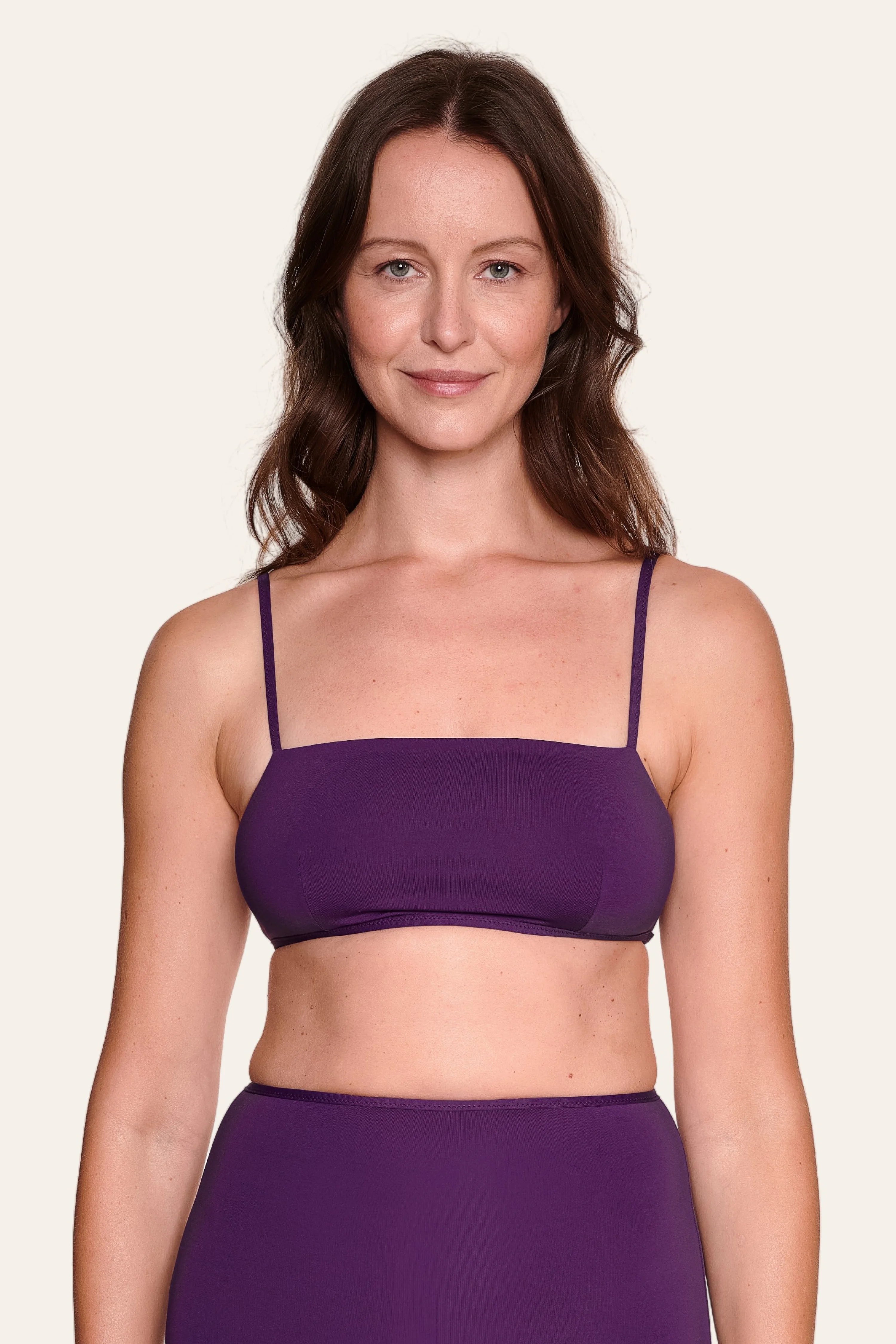 Frau trägt MYMARINI Bikinitop mit Spaghettiträgern in Lila, lächelnd vor neutralem Hintergrund.#color_lavender-plum