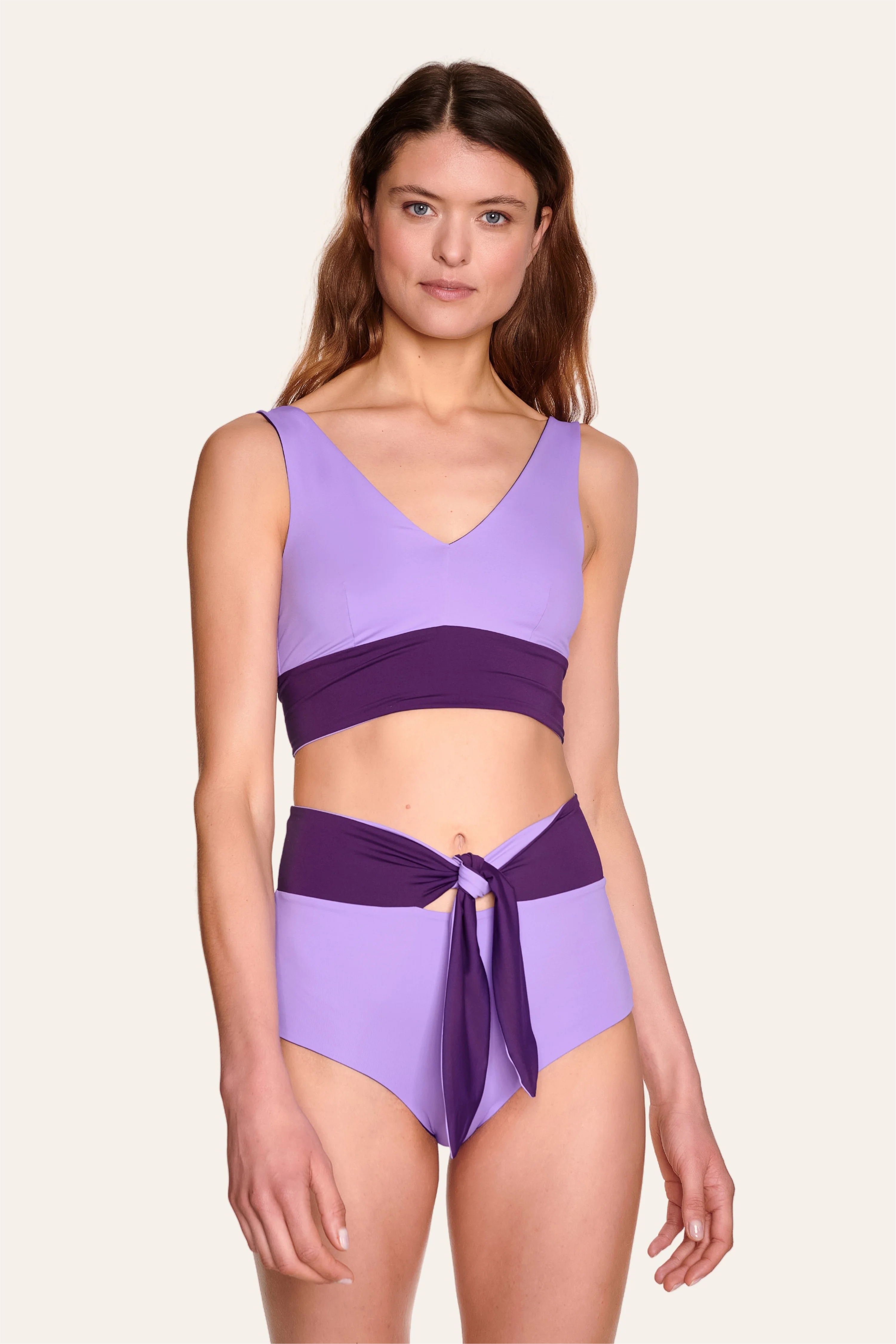 Frau trägt MYMARINI Bikini mit V-Ausschnitt und High Waist Schnitt in Lila und Dunkelviolett vor hellem Hintergrund.#color_lavender-plum