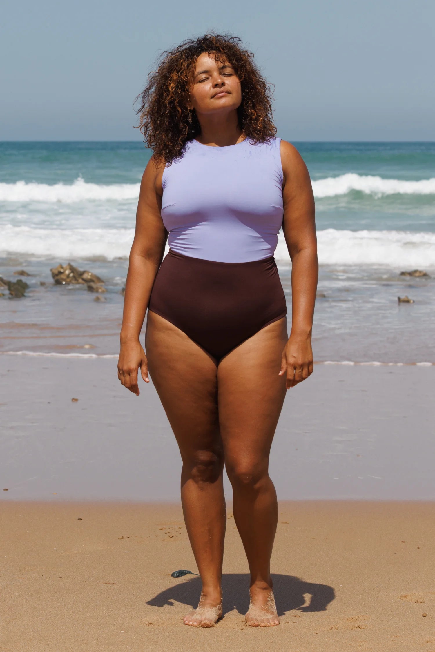 Frau am Strand in MYMARINI Onepiece Badeanzug lilafarben und braun; Blick zur Sonne, Meer im Hintergrund.#color_chocolate-lilac