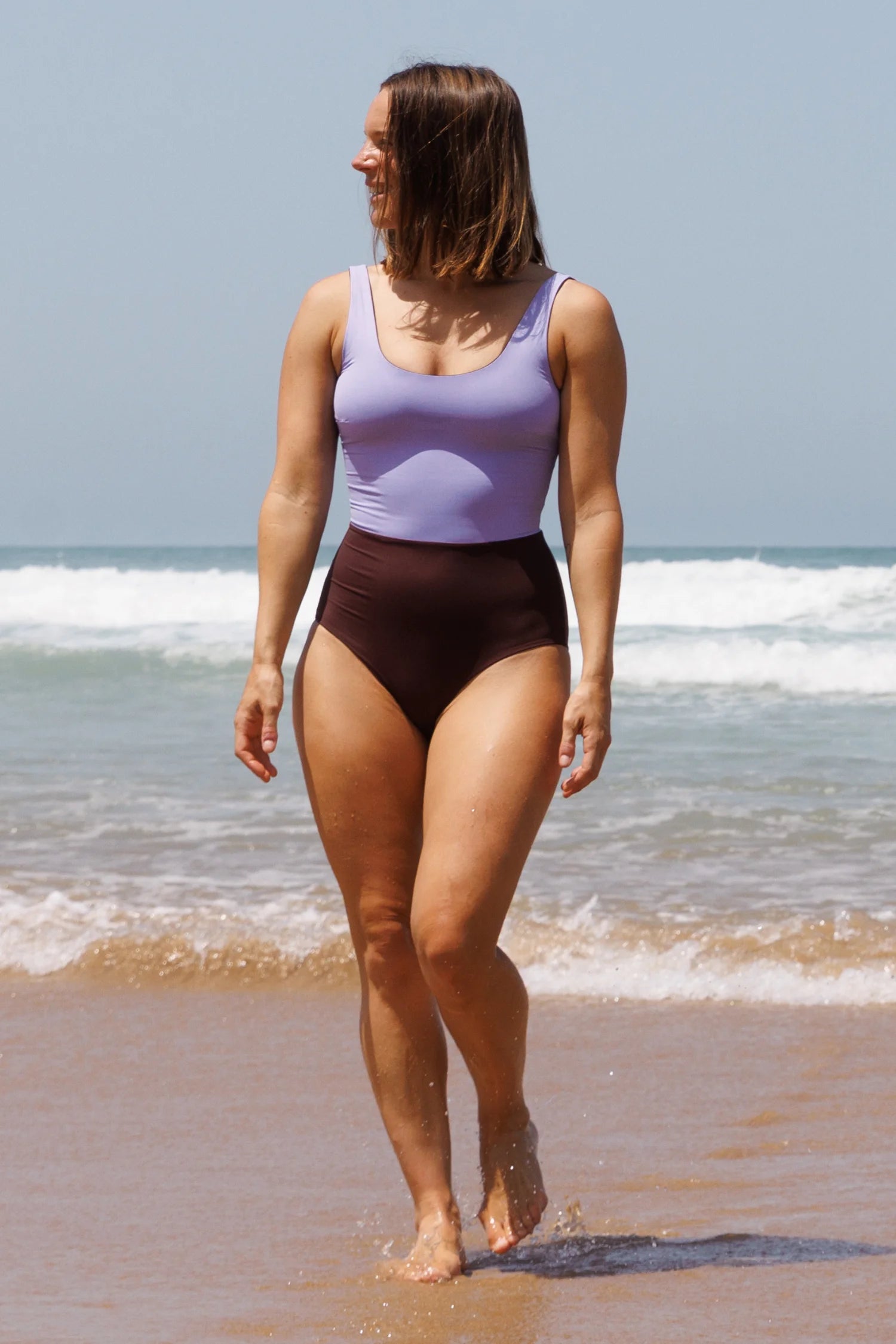MYMARINI Bikinitop in Flieder mit Breite Trägern und braune High-Waist Bikinihose; Sandstrand, Wasser im Hintergrund, Person geht entlang.#color_chocolate-lilac