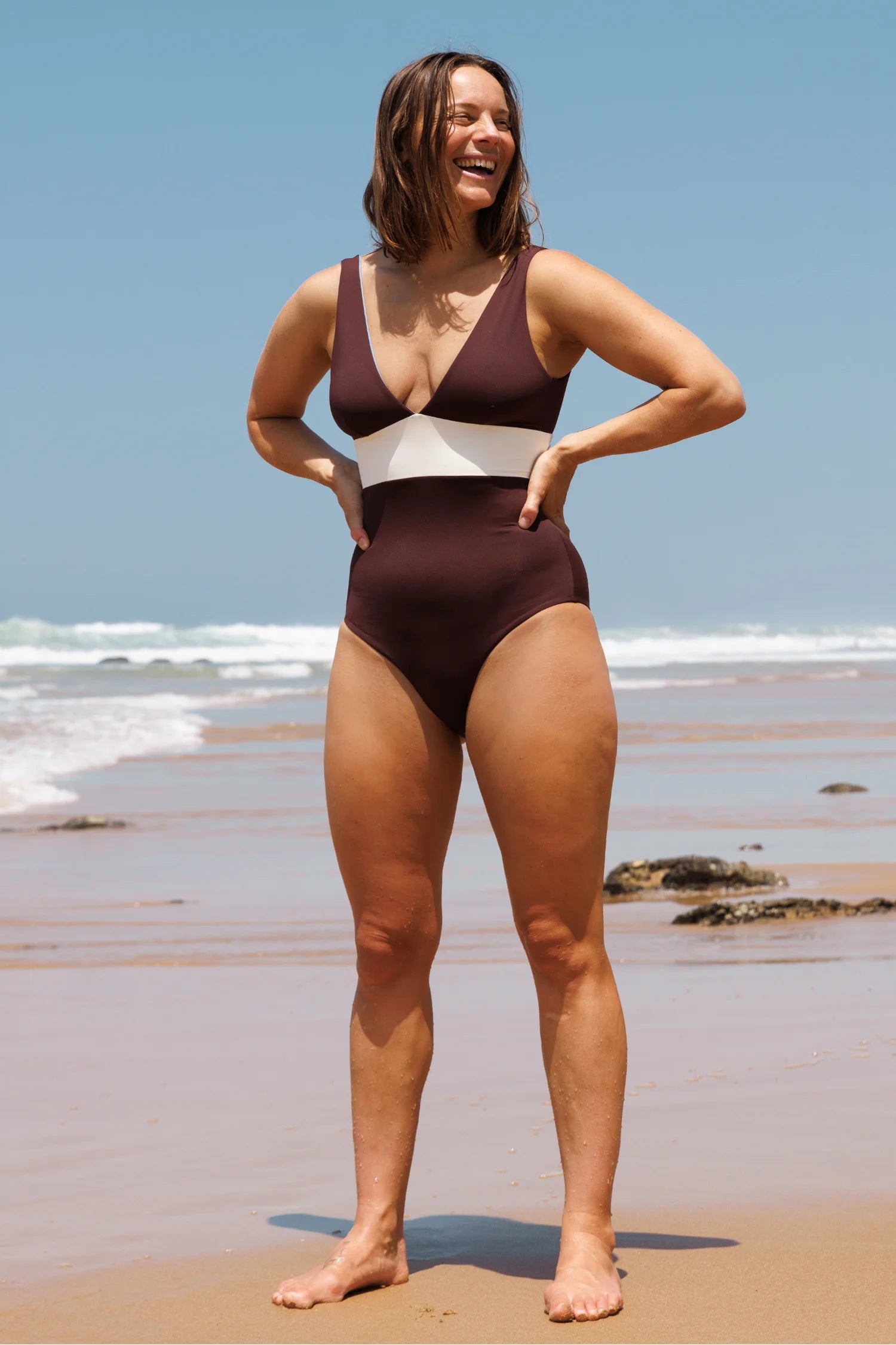 MYMARINI Badeanzug in braun mit weißer Mittelbinde, V-Ausschnitt, hoher Beinausschnitt, am Strand stehend, lächelnd.#color_chocolate-lilac