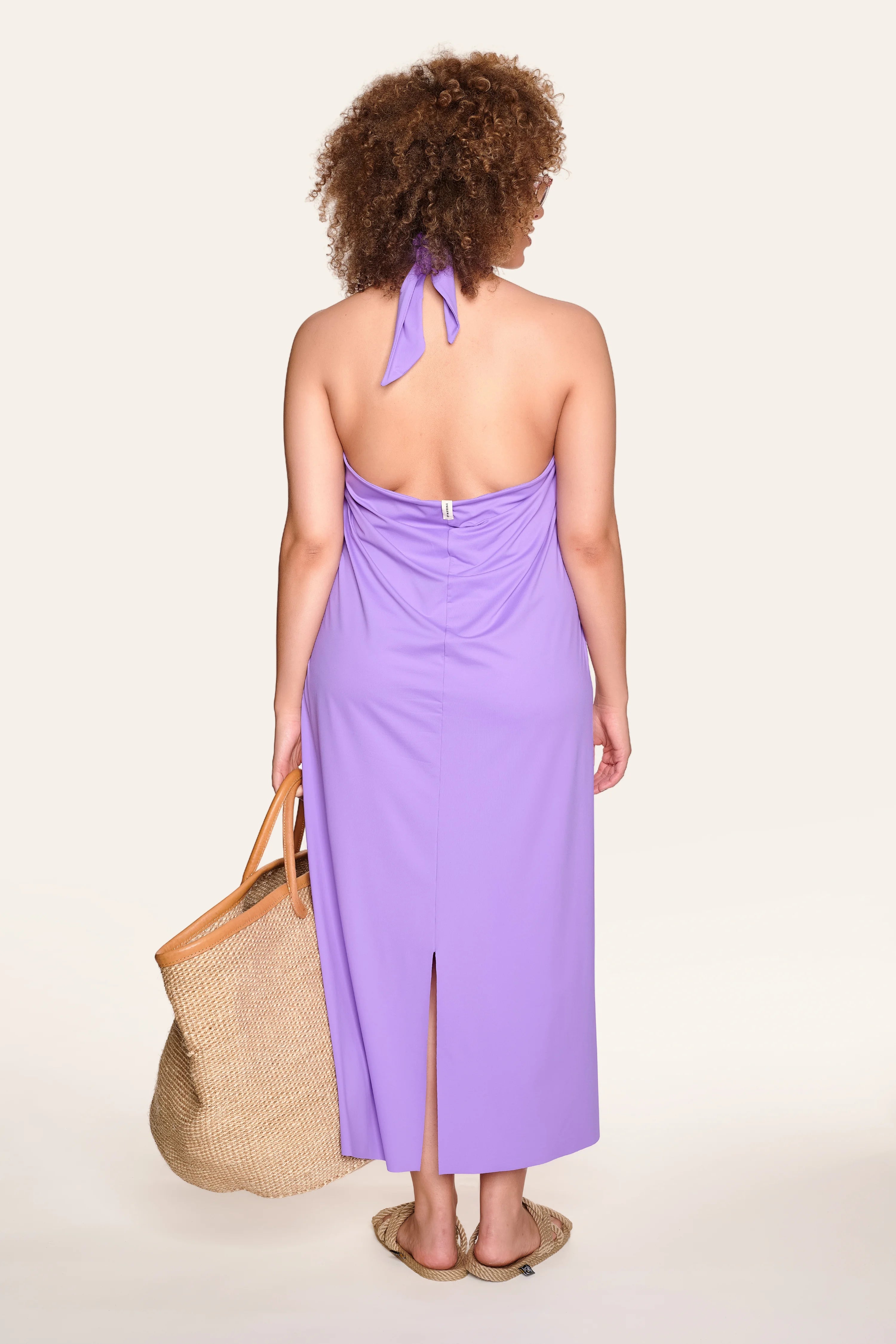 Person von hinten in lila MYMARINI-Kleid mit Neckholder, trägt Strohtasche und Sandalen.#color_lavender