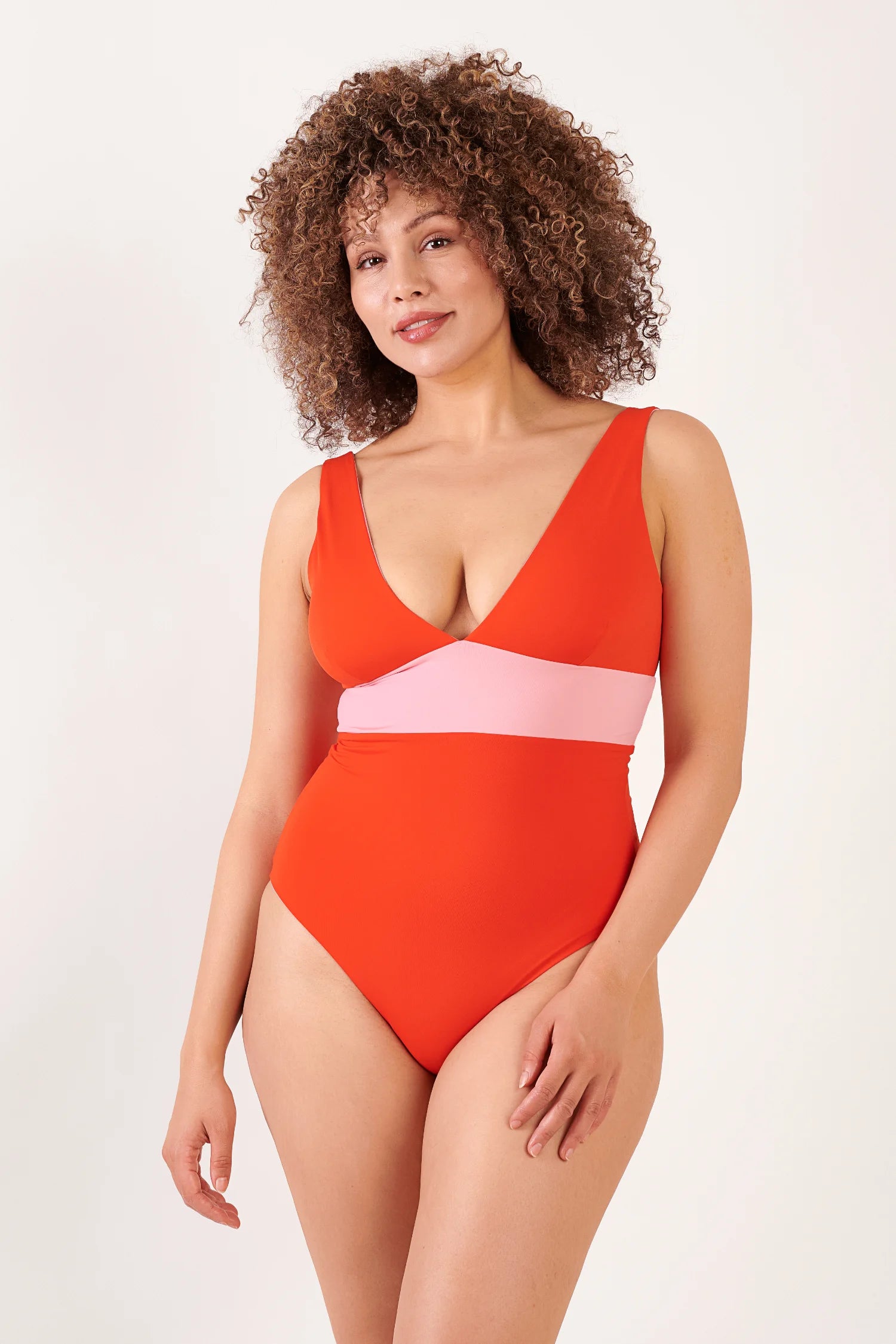 MYMARINI One-Piece-Badeanzug in Rot mit rosa Band unter der Brust, V-Ausschnitt, breite Träger – Frau mit lockigem Haar.#color_poppy-macaron