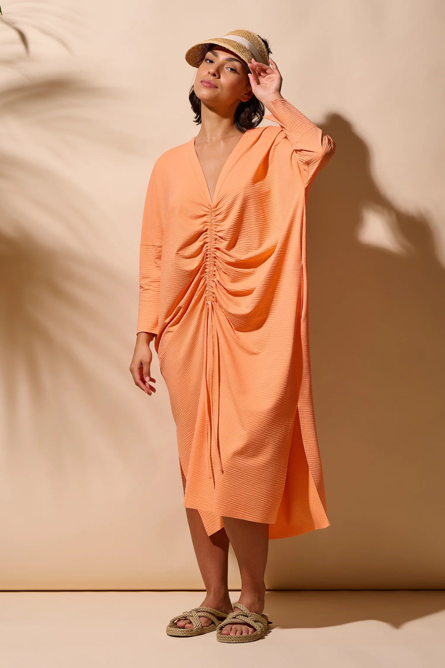 Person trägt ein MYMARINI-Kleid in Orange mit V-Ausschnitt und langen Ärmeln, steht vor beigem Hintergrund.#color_peach