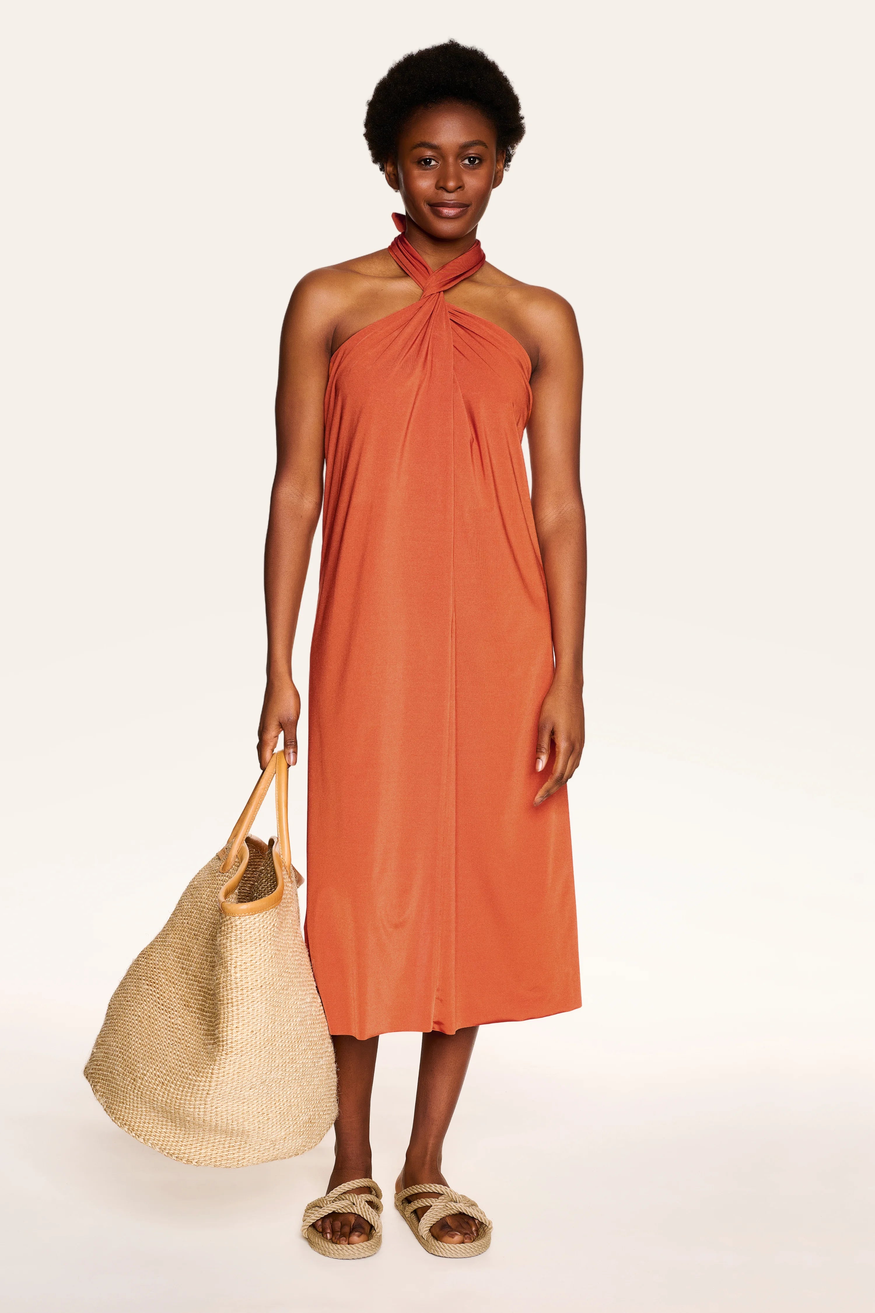 Person in orangefarbenem MYMARINI-Kleid mit Neckholder-Schnitt, hält eine große Strohtasche, trägt Sandalen.#color_papaya