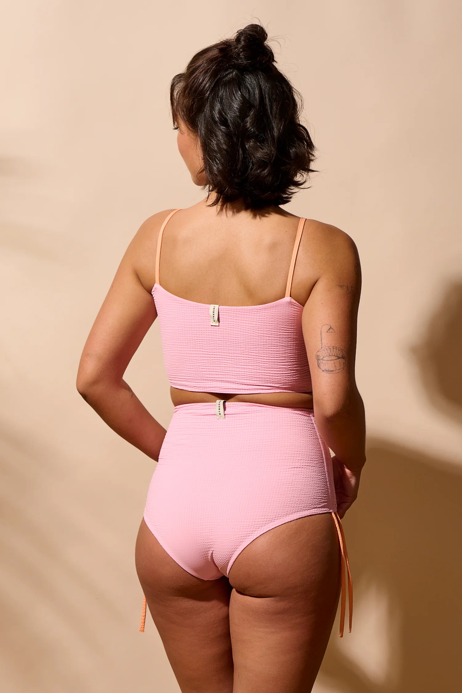 Frau in rosa MYMARINI-Bikini mit Bandeau-Top und High-Waist-Hose, von hinten vor beigem Hintergrund.#color_marshmallow-peach