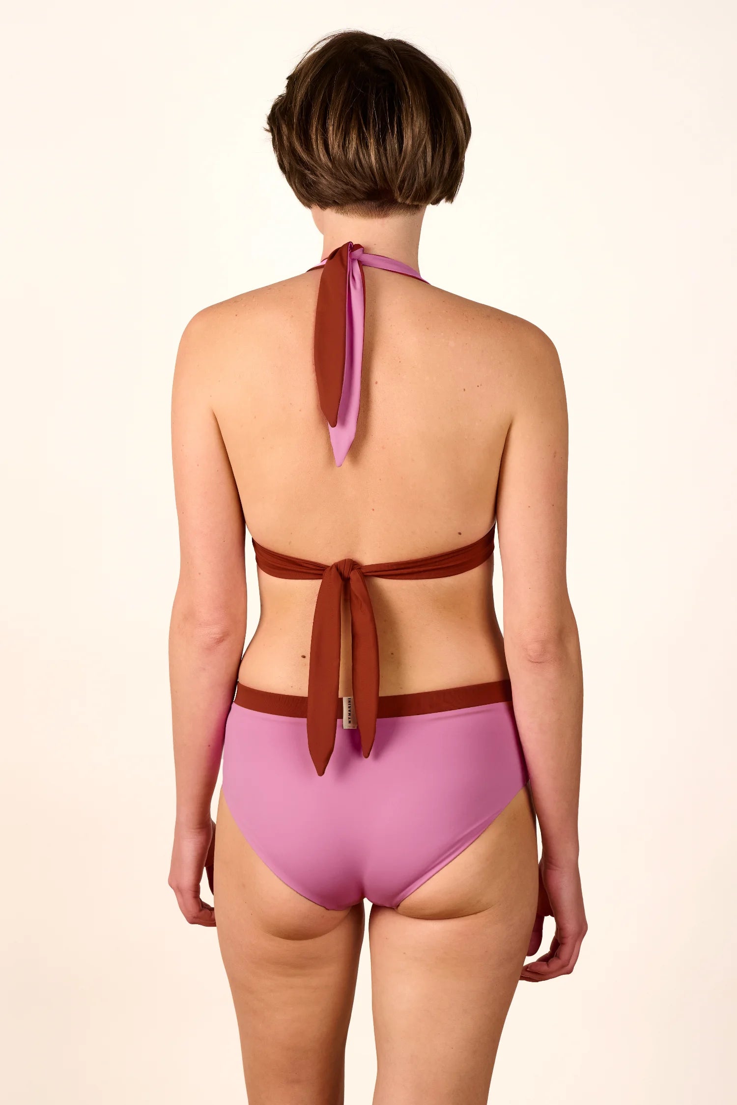 Frau in MYMARINI Bikinihose und Neckholder-Top, Rückansicht, hoher Beinausschnitt, rosa und braun, vor hellem Hintergrund.#color_sienna-orchid