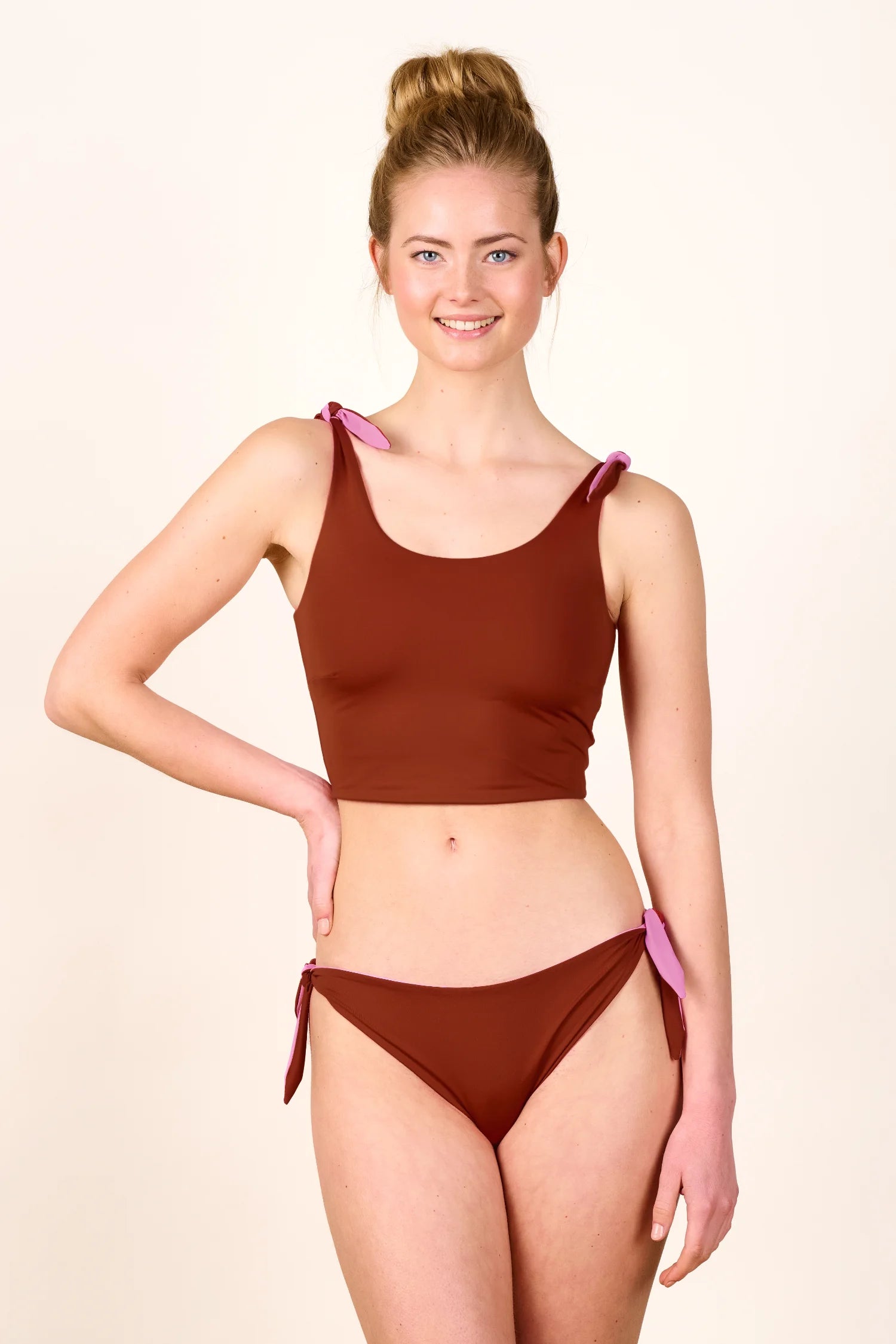 Frau trägt MYMARINI Bikini mit breiten Trägern und Mid rise Schnitt in Braun, lächelnd vor neutralem Hintergrund.#color_sienna-orchid