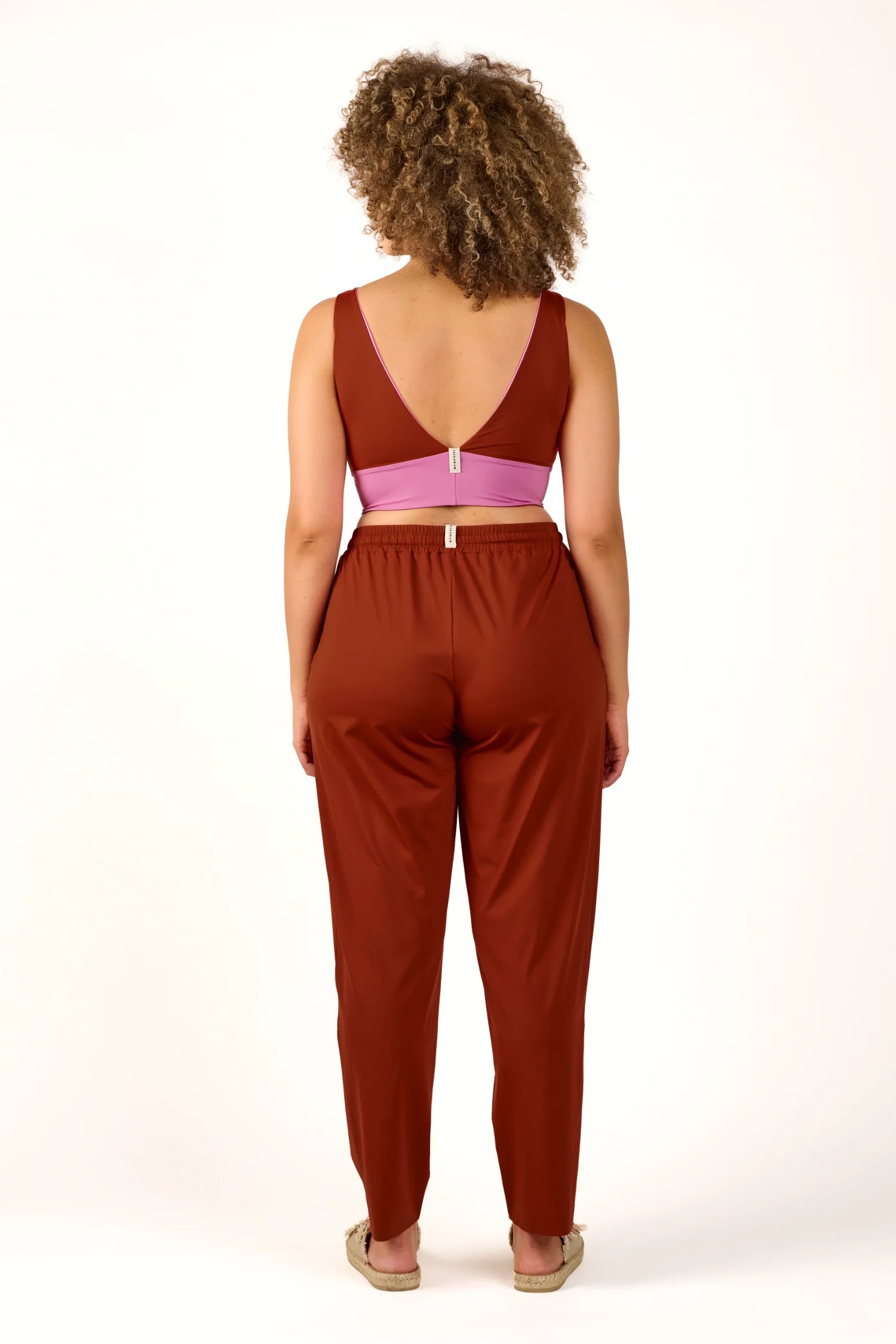Person von hinten in MYMARINI Badeanzug mit V-Ausschnitt und hoher Taille, kombiniert mit brauner Hose.#color_sienna