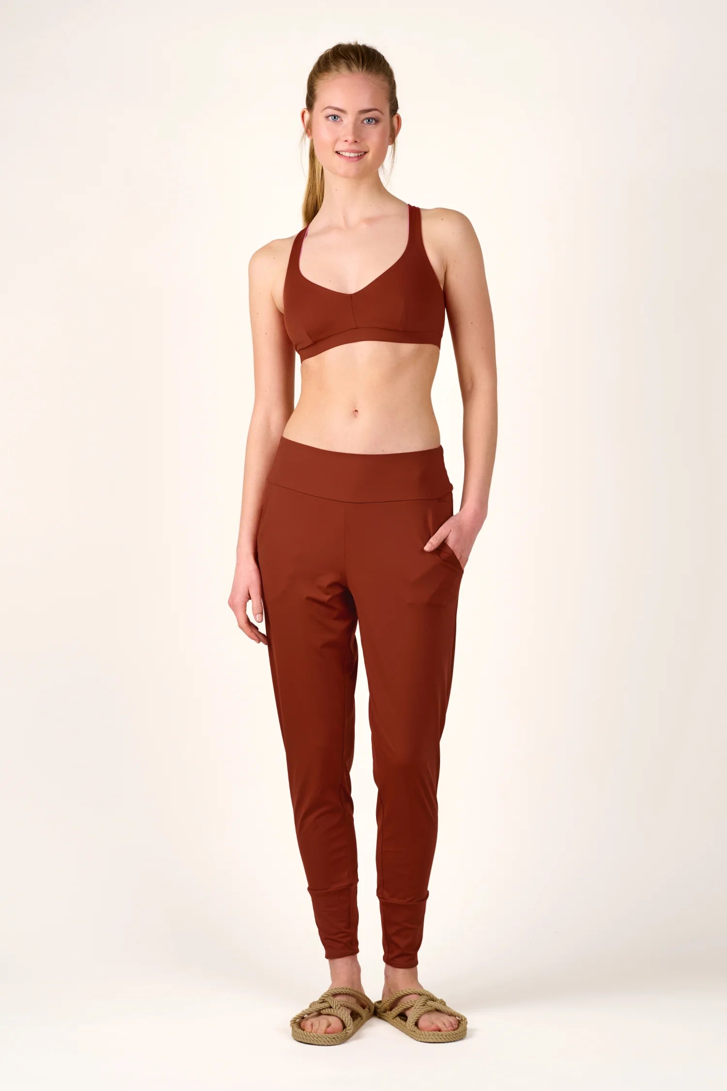 Frau trägt MYMARINI Bikinitop mit V-Ausschnitt und Hose in Braun, stehend vor neutralem Hintergrund, lächelnd.#color_sienna