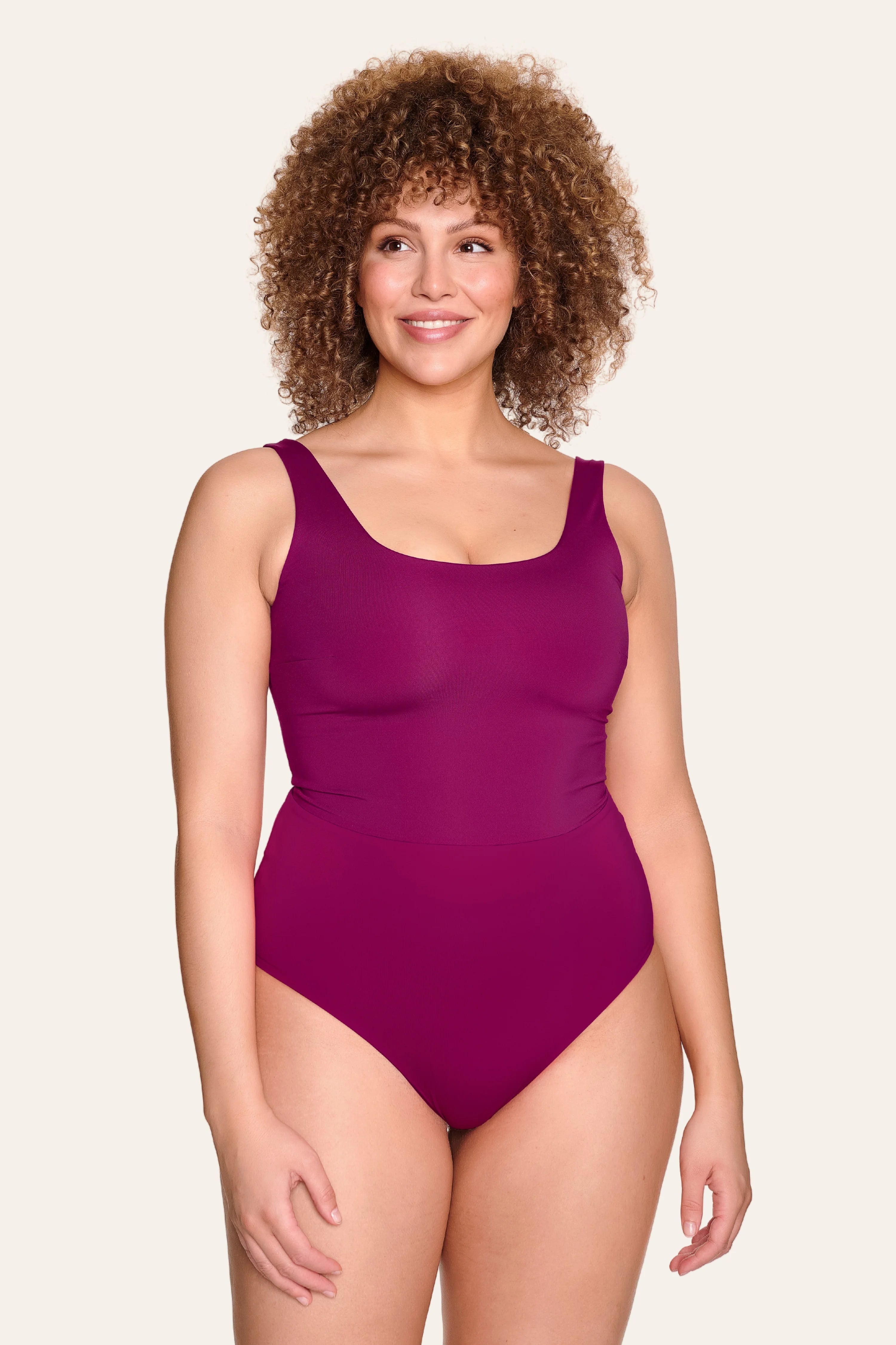 Frau trägt einen MYMARINI Badeanzug in Lila mit breiten Trägern und hohem Beinausschnitt, lächelnd vor hellem Hintergrund.#color_pink-hortensia