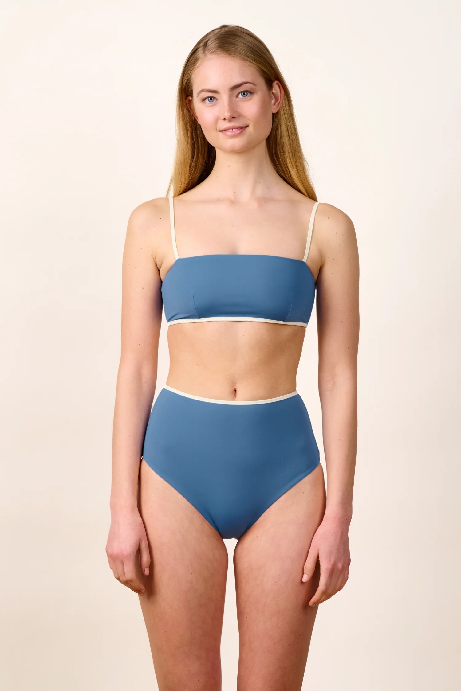 Frau trägt MYMARINI Bikinitop mit Spaghettiträgern und High-Waist-Bikinihose in Blau vor neutralem Hintergrund.#color_coast-desert
