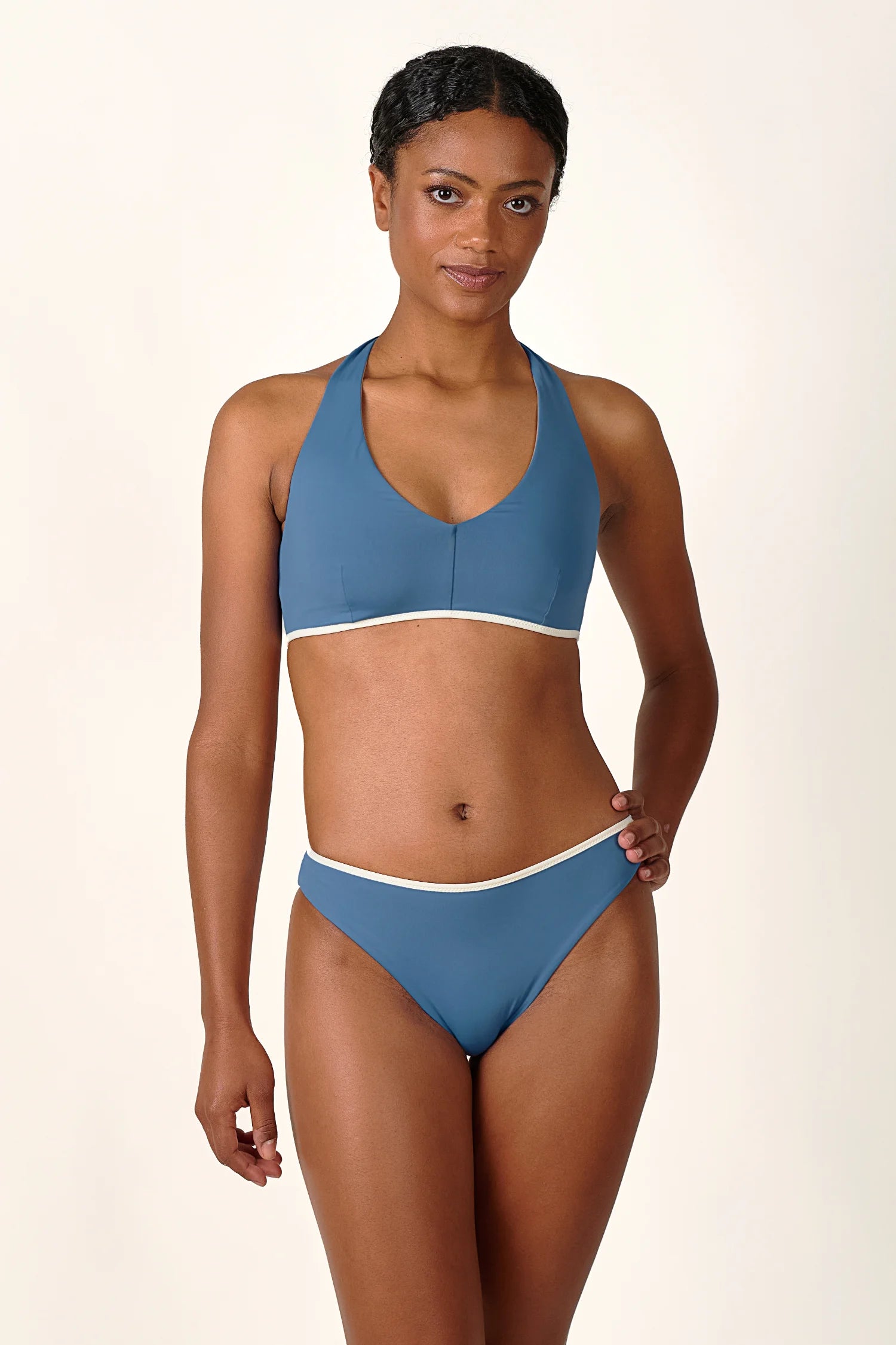 Frau trägt MYMARINI Bikini mit Neckholder-Top und Mid-rise-Hose in Blau vor hellem Hintergrund.#color_coast-desert