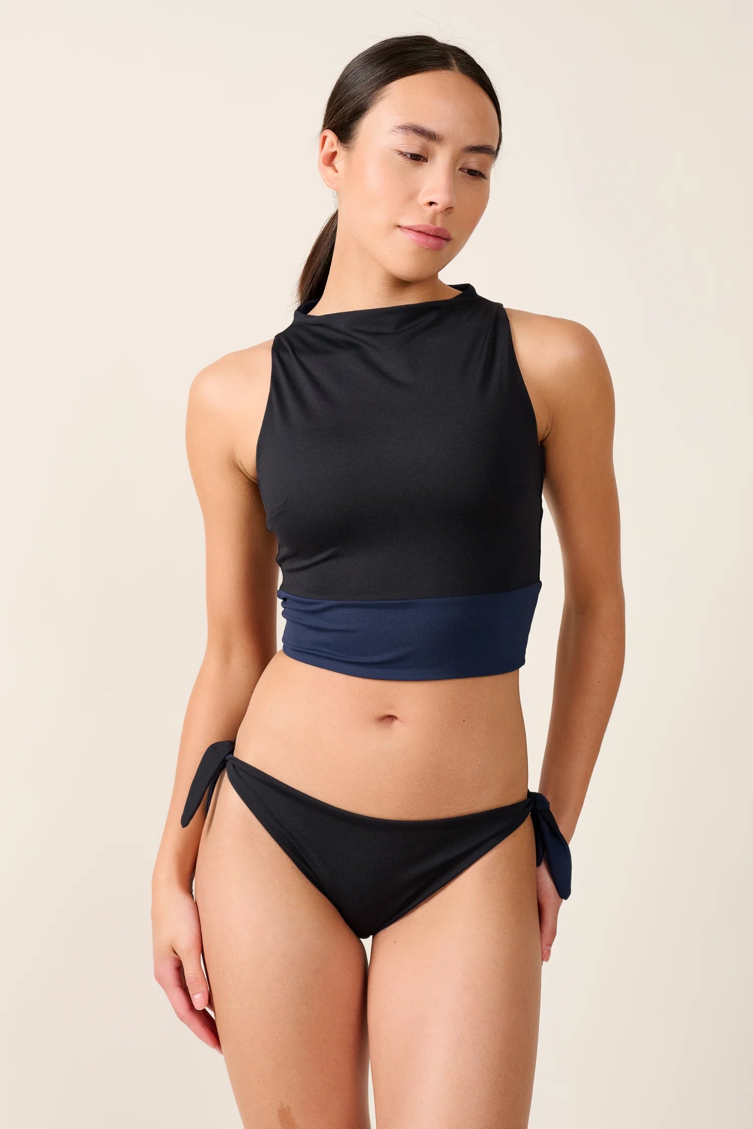 Frau trägt MYMARINI Bikinihose mit seitlichen Schleifen und hochgeschlossenem Top in Schwarz und Blau vor neutralem Hintergrund.#color_black-navy