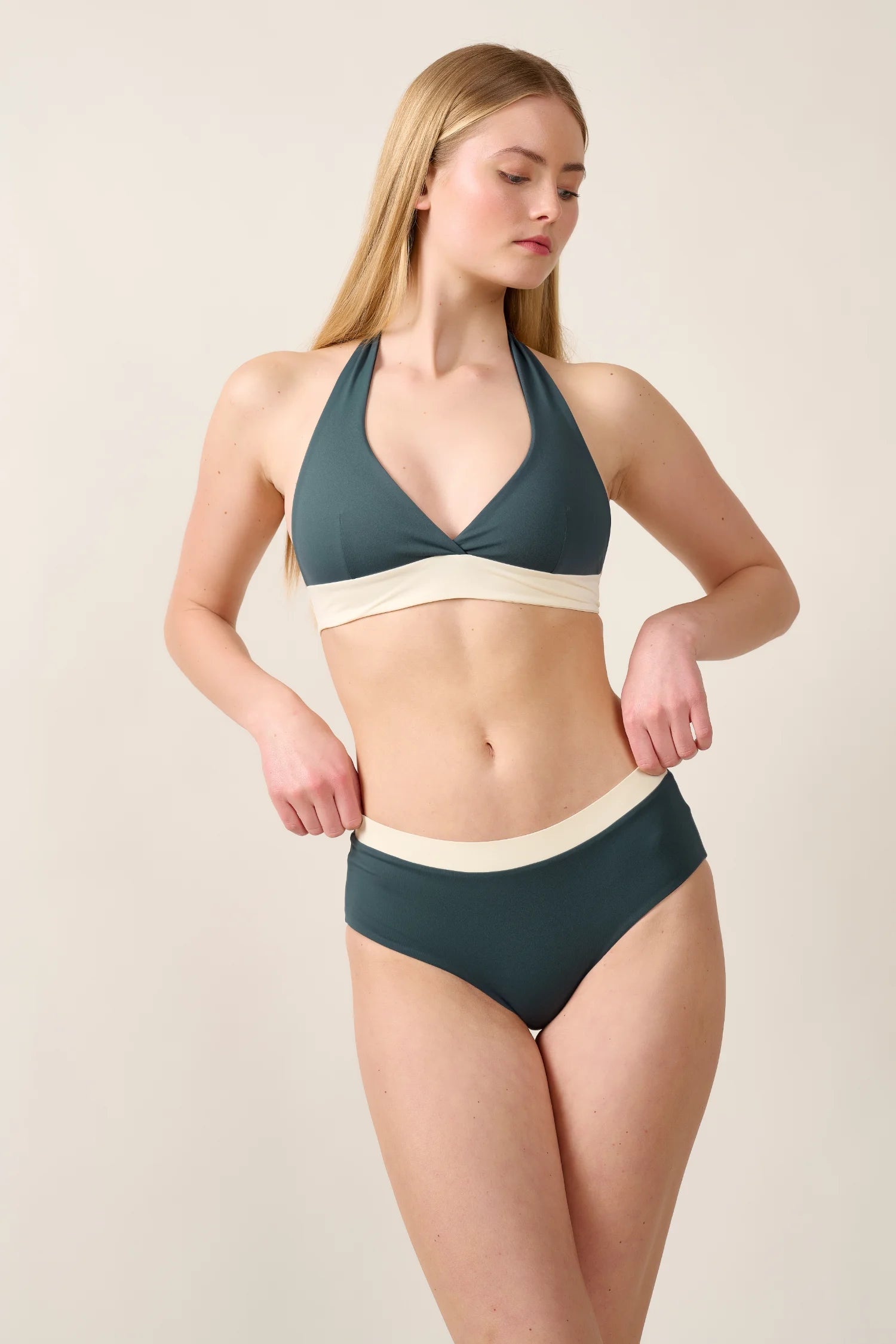 Frau trägt MYMARINI Bikini mit Neckholder-Top und High-Waist-Hose in Dunkelgrün und Creme vor neutralem Hintergrund.#color_black-grey-white