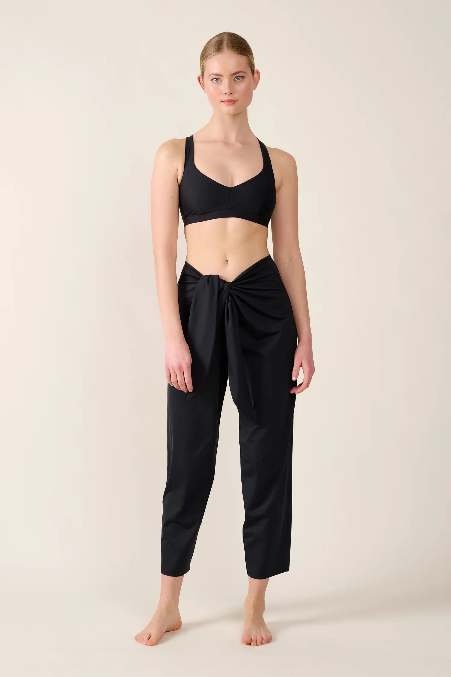 Frau trägt MYMARINI Bikinitop mit breiten Trägern und Hose. Elegante, schlichte Präsentation vor neutralem Hintergrund.#color_black