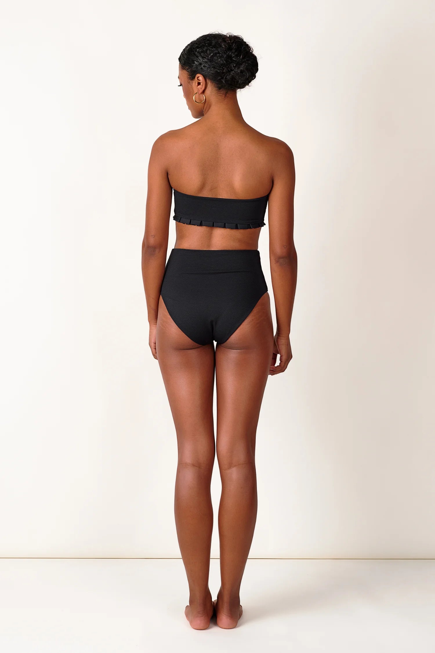 Frau in schwarzem MYMARINI Bandeau-Bikinitop und High-Waist-Bikinihose, von hinten, vor hellem Hintergrund.#color_black